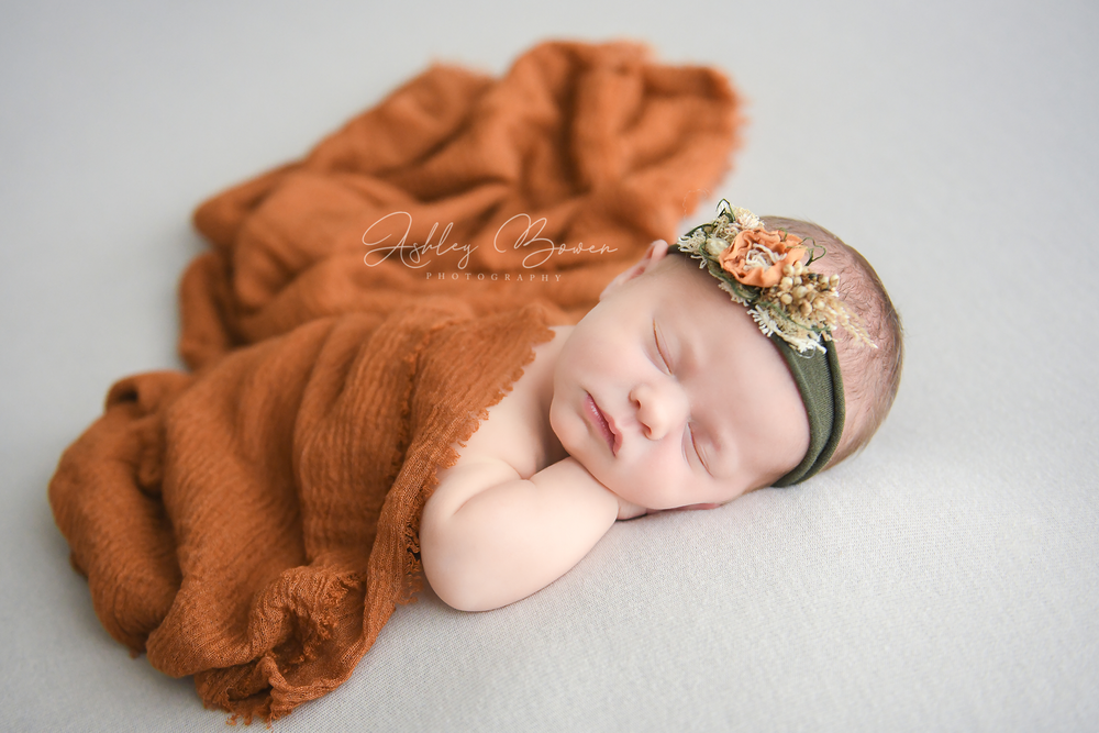 Sleeping newborn baby wrapped in rust orange knit blanket with floral headband on white backdrop.