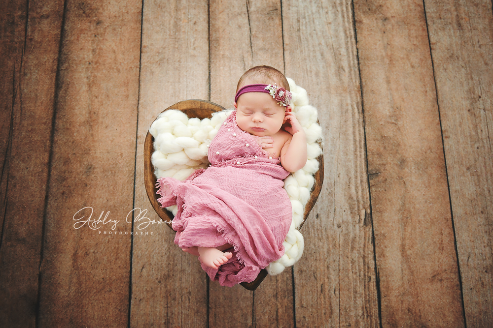Newborn baby girl in pink wrap sleeping in heart-shaped wooden bowl with white yarn.