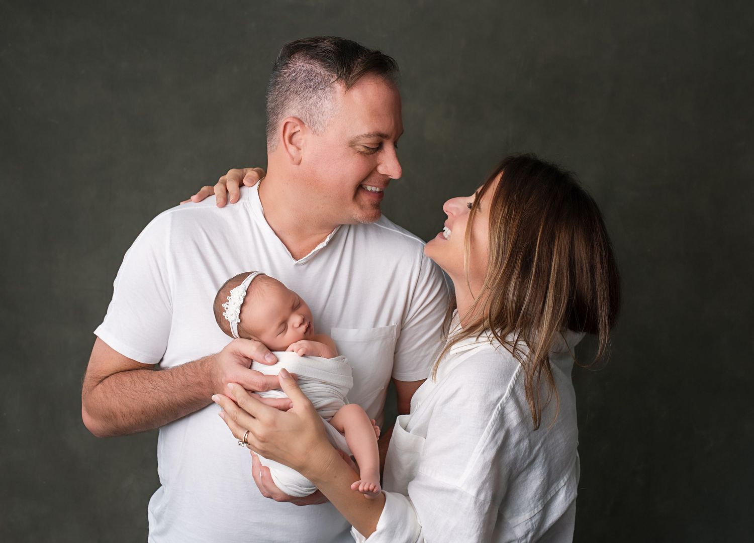 Happy parents in white shirts smile at their newborn baby during a professional studio portrait session.