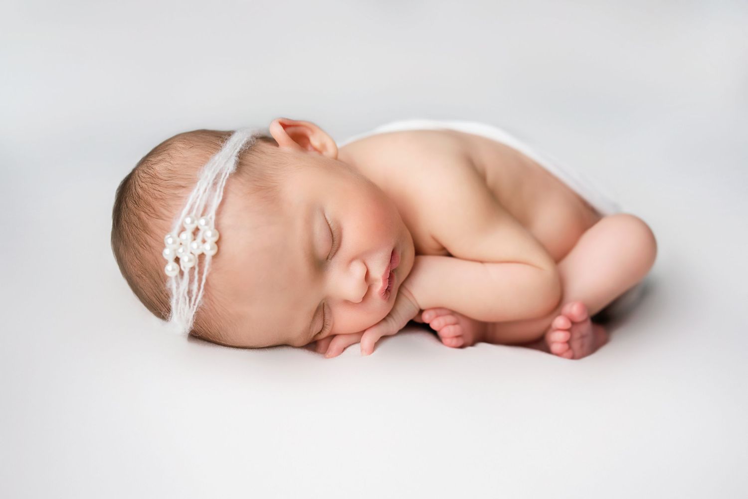 Smiling sleeping newborn baby girl with white floral headband curled up on soft white blanket.