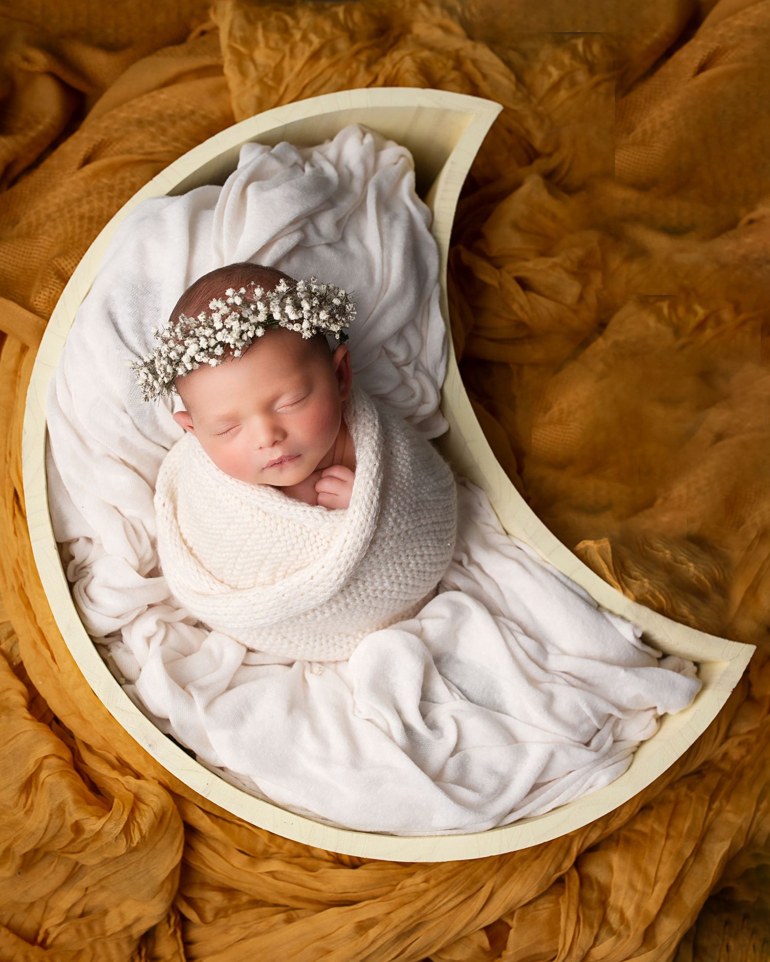 Sleeping newborn baby wrapped in cream knit swaddle in moon prop with floral crown for studio photo.