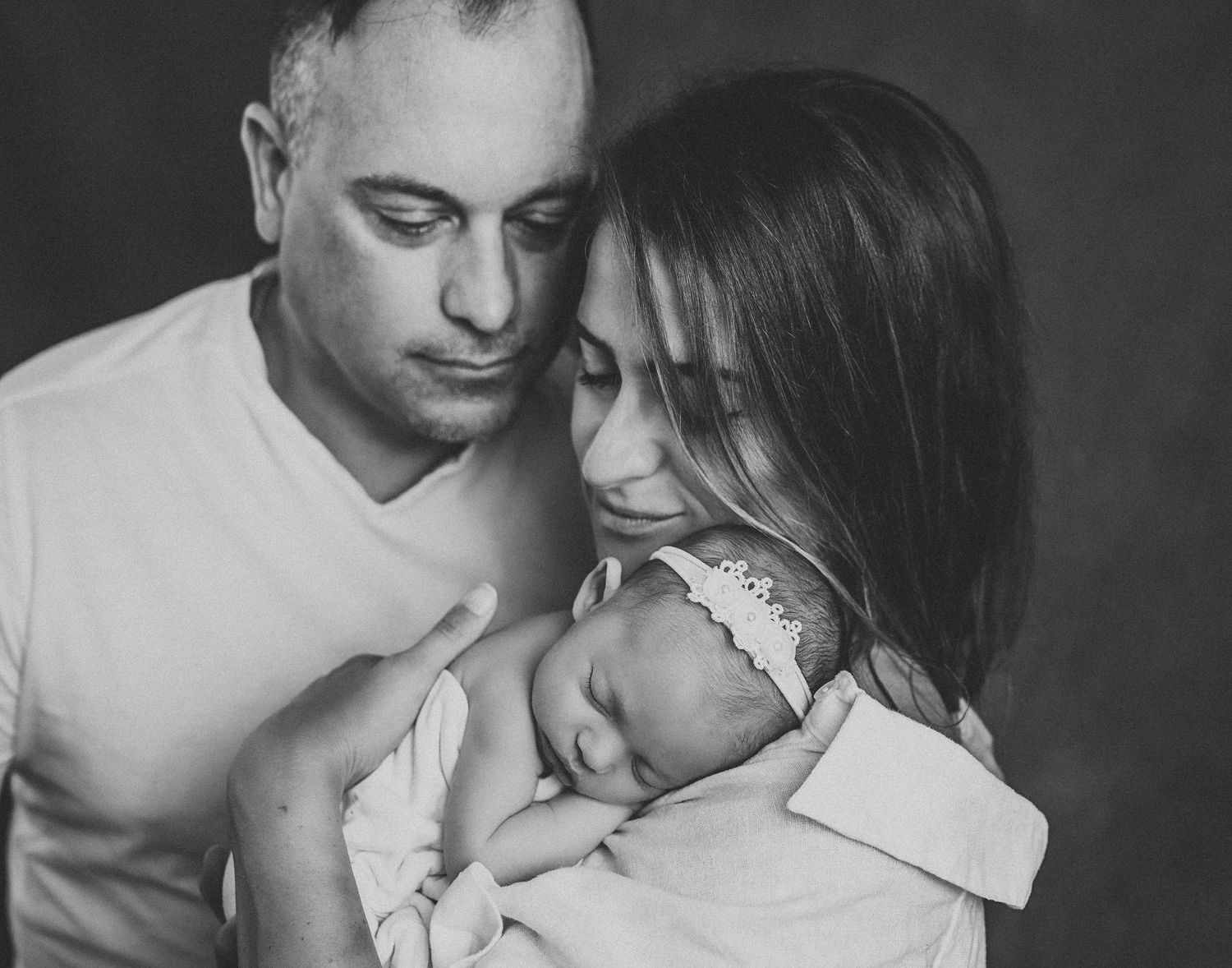 Black and white portrait of parents lovingly gazing at their sleeping newborn baby together.