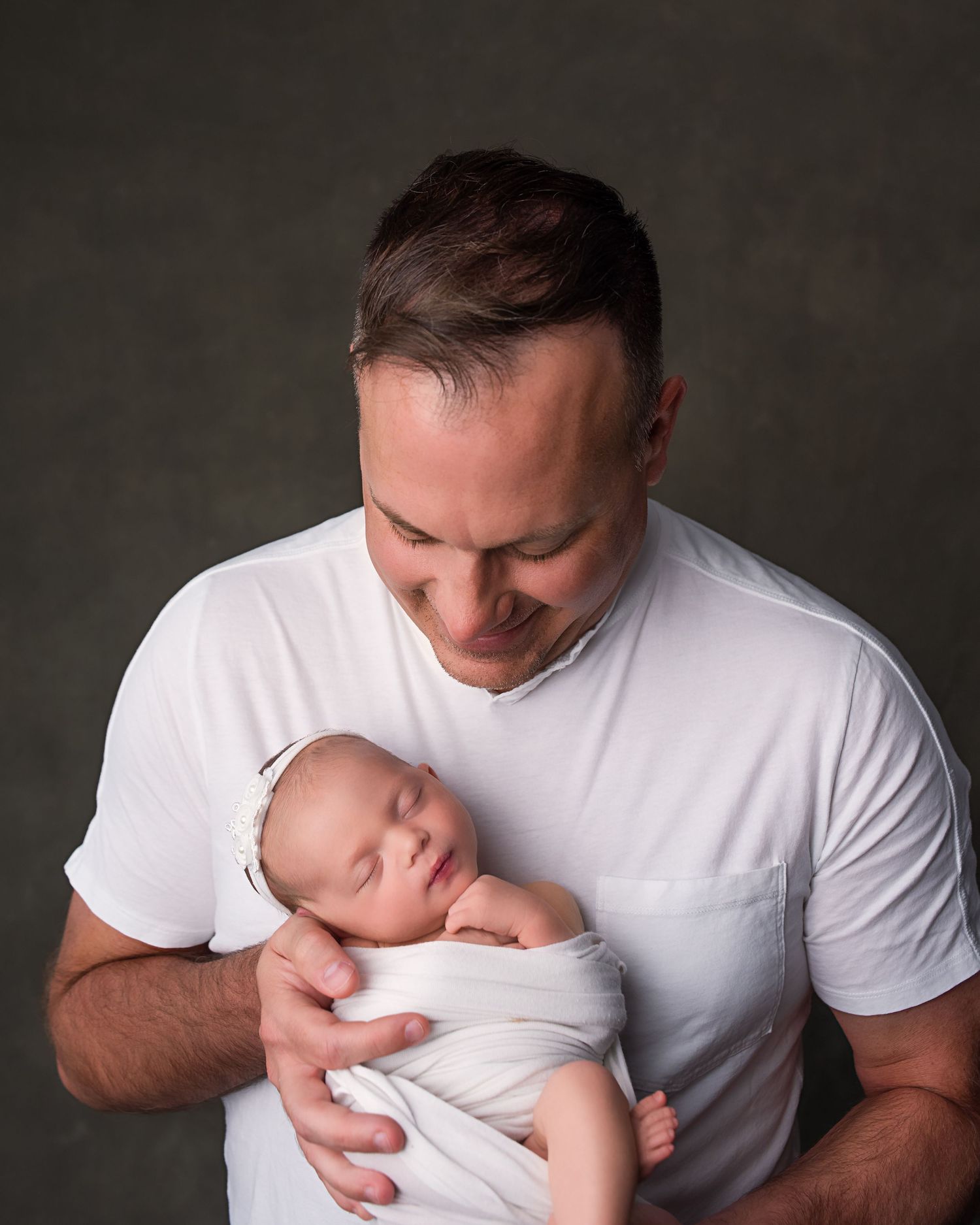 Proud father in white t-shirt smiling down at sleeping swaddled newborn baby in studio portrait.
