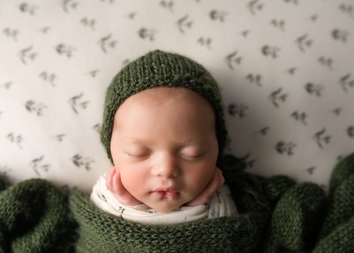 Sleeping newborn baby in green knitted bonnet and matching wrap on floral patterned white blanket.