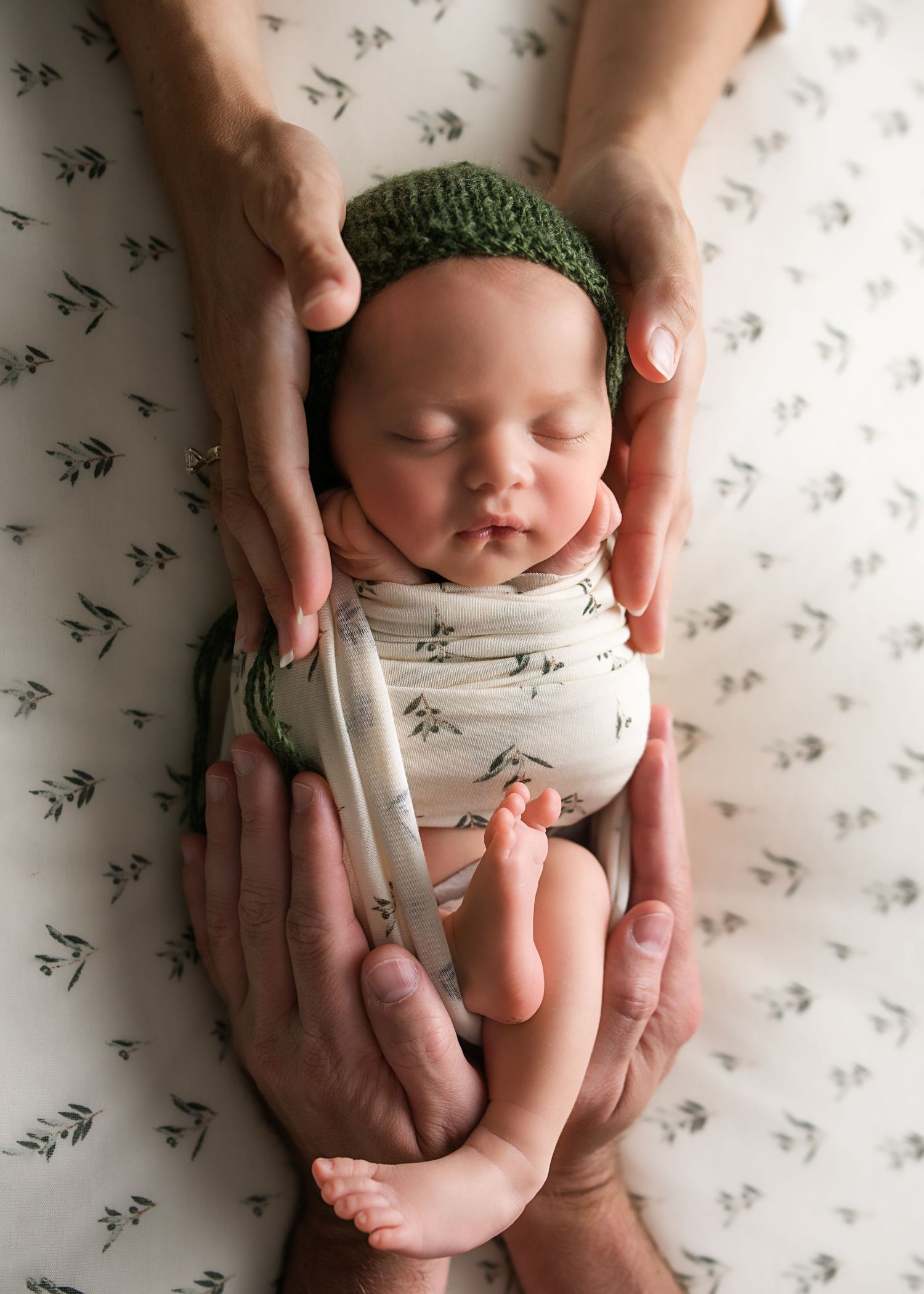 Parents gently cradling swaddled newborn in green bonnet on floral blanket in professional newborn photo session.