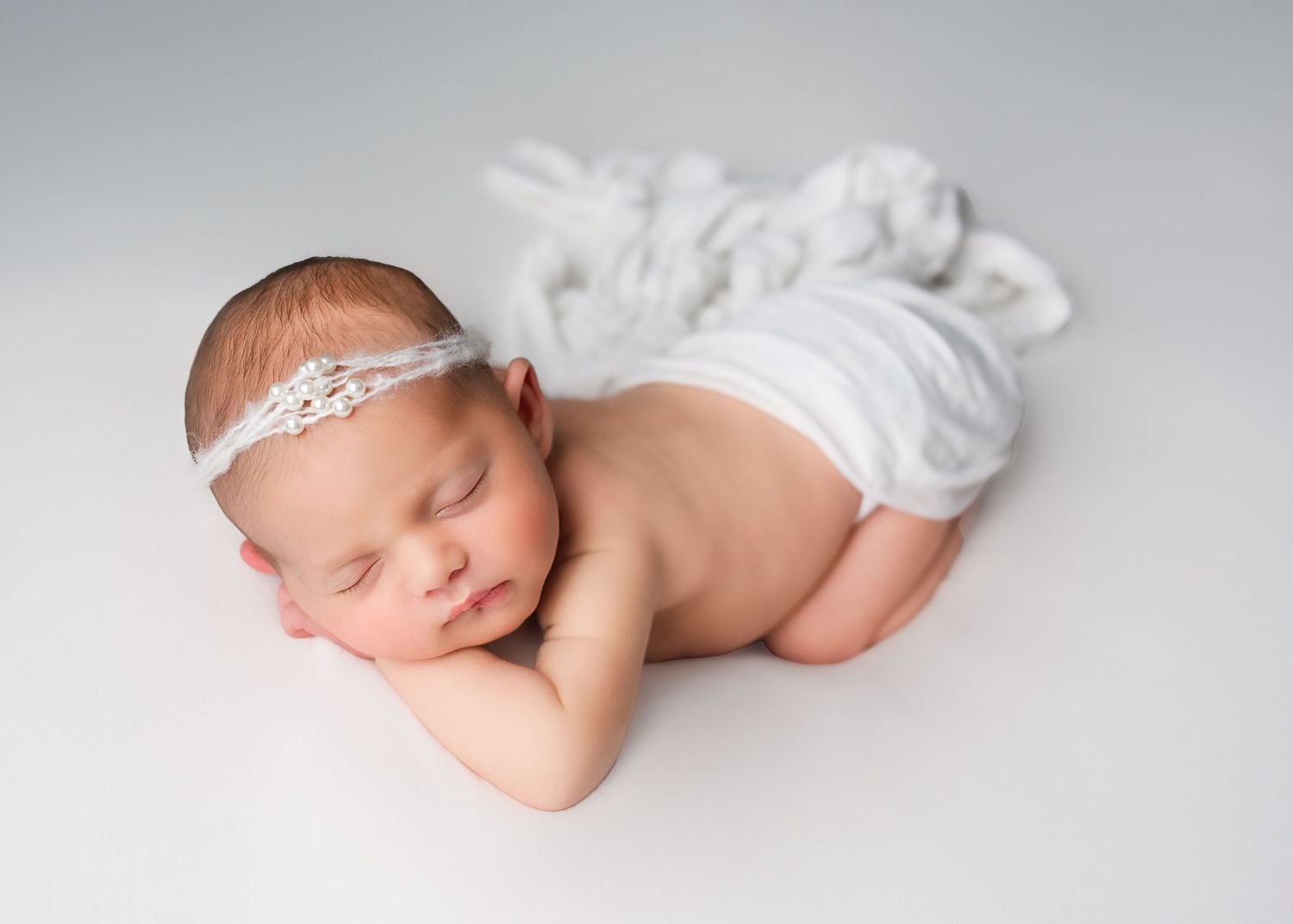 Peaceful newborn baby girl with lace headband posed on white background with soft draped fabric.