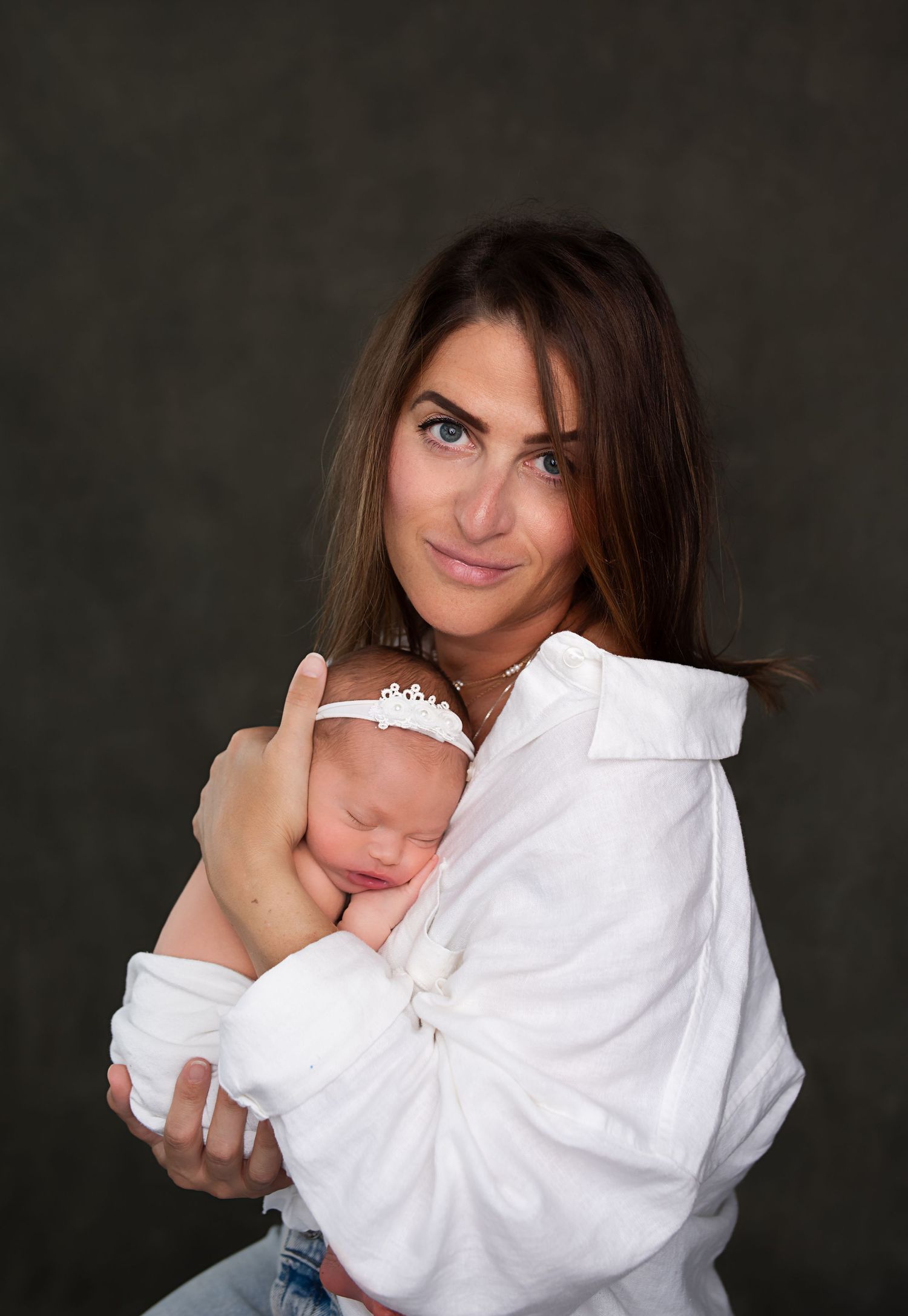 Brunette mother in white shirt tenderly holding sleeping newborn baby against dark studio background.
