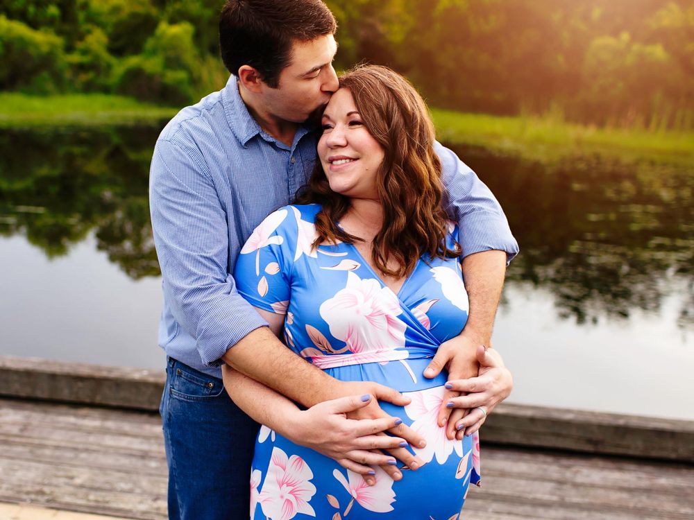 Pregnant wife being hugged from behind by husband while holding belly in Alvin Tx