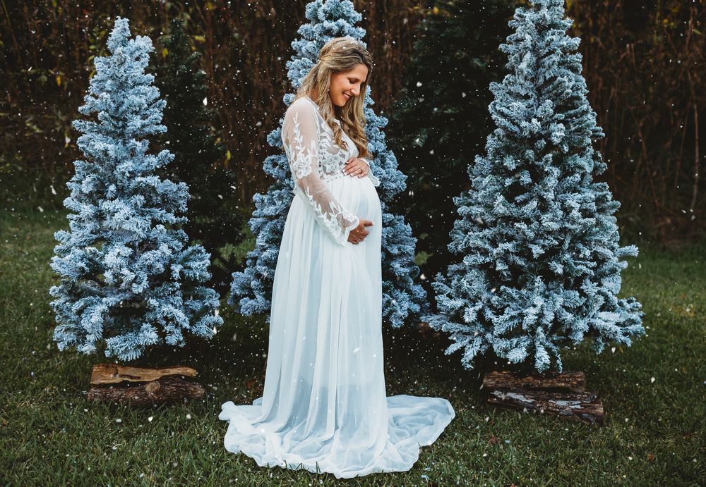 Mama ini white maternity gown holding belly surrounded by Christmas trees in Alvin Tx