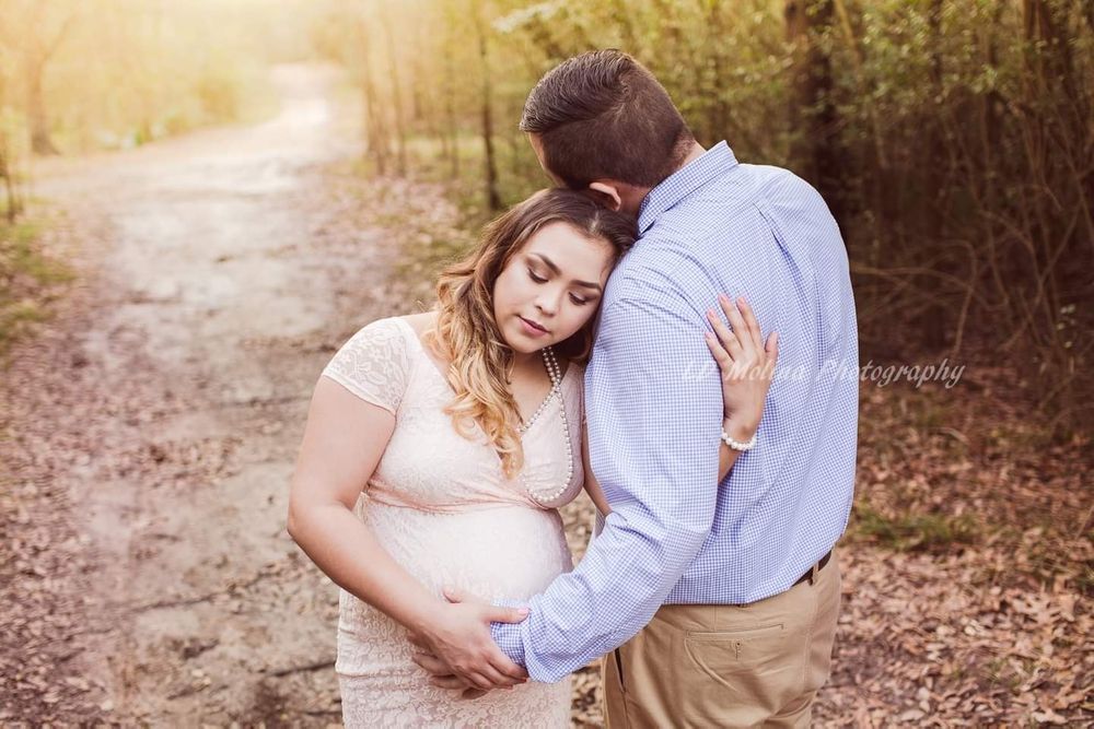 Couple embracing on forest path during sunset with pregnant woman in white dress and man in blue shirt.