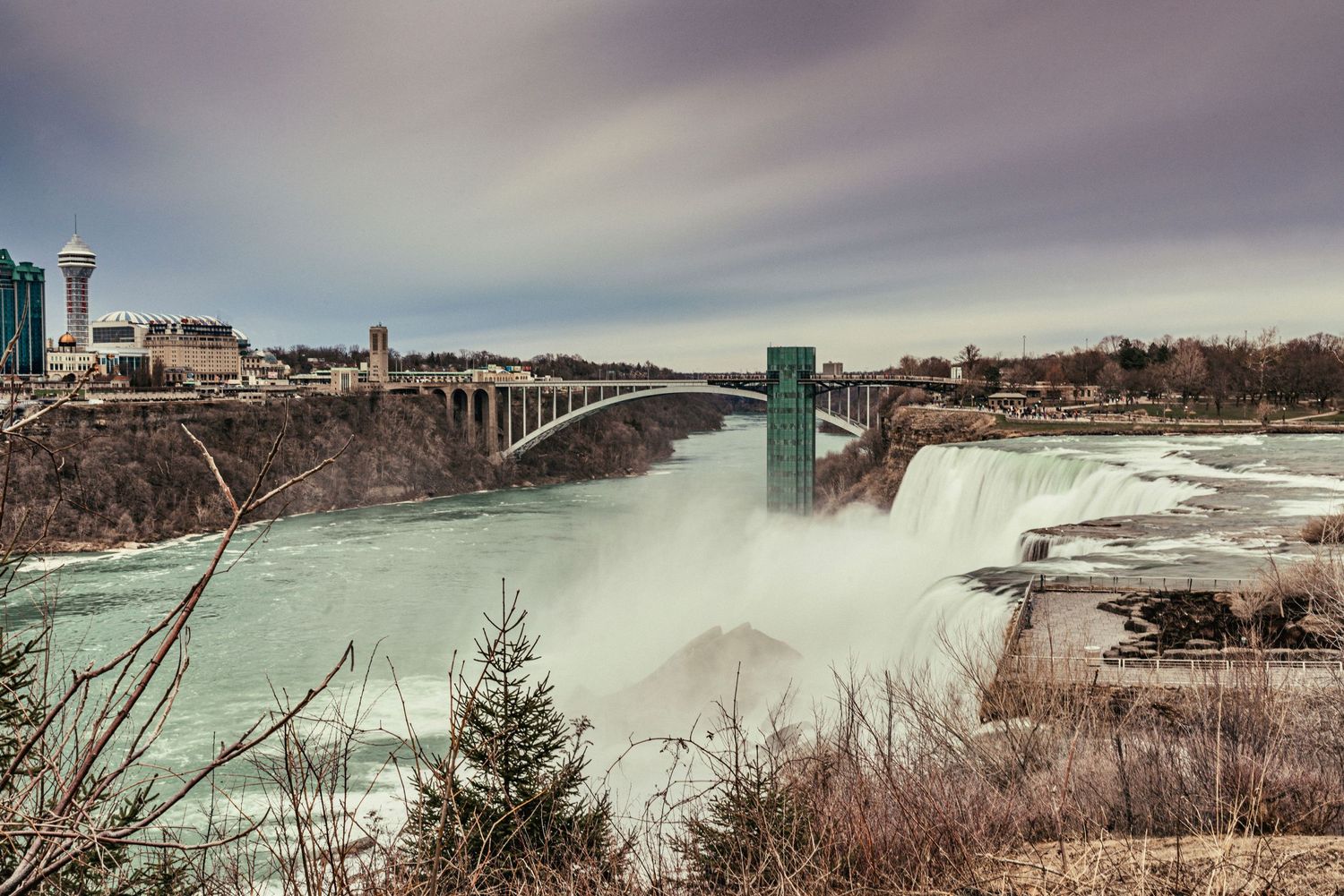 Wide view of Niagara Falls with observation deck and bridge visible in winter landscape.