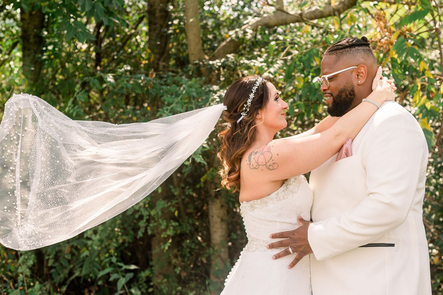 Elegant wedding couple embracing outdoors with flowing veil and natural greenery backdrop.