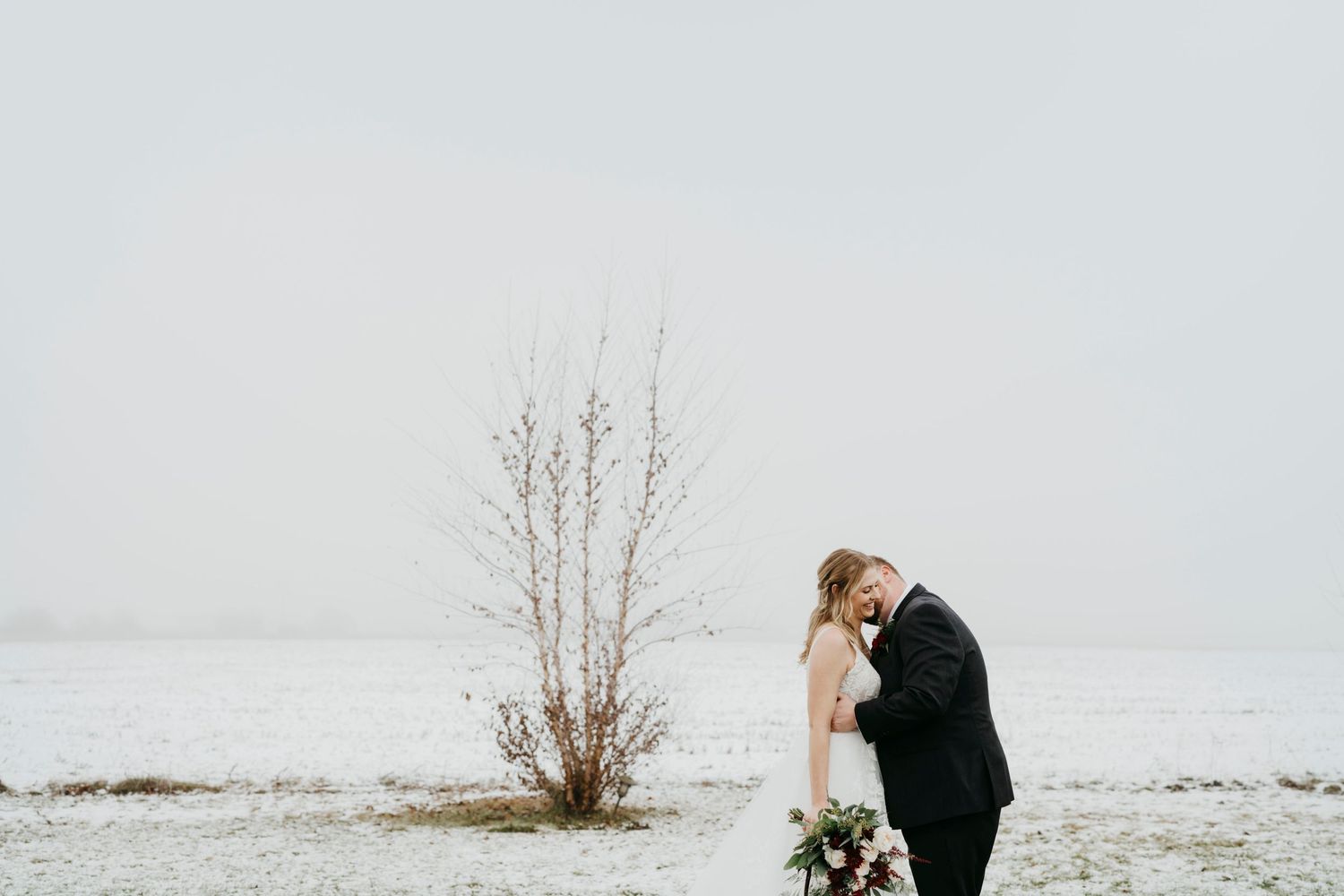 Winter wedding portrait in a minimalist snowy landscape with bare trees.