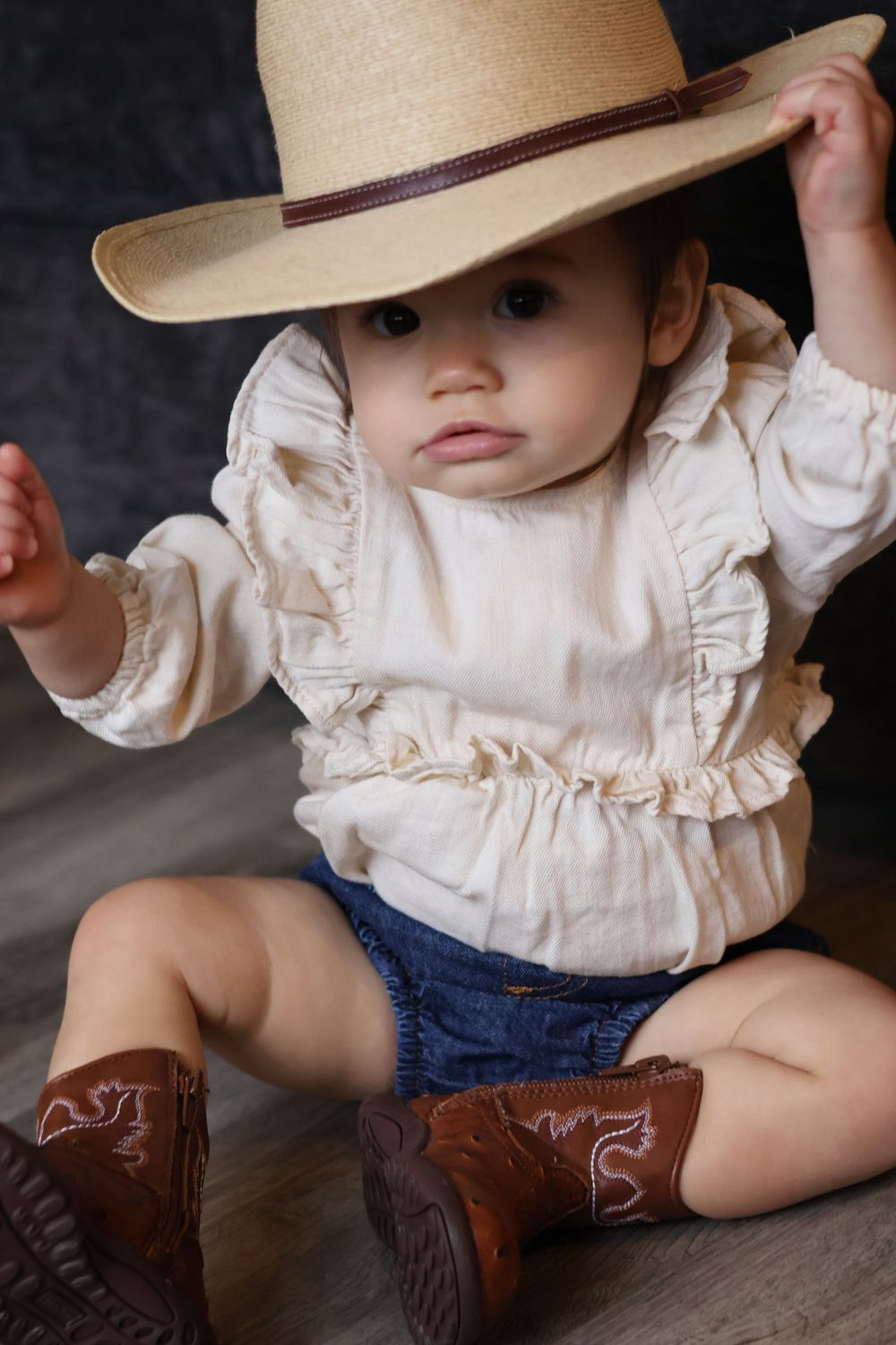 Small child wearing a cowboy hat and western boots sits on dark background.