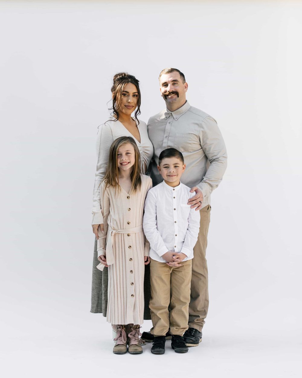 A happy family of four poses together, parents with two children in neutral tones against a white background.