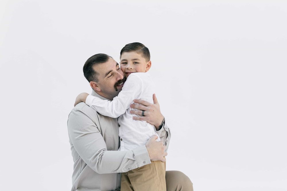 A father joyfully hugs his young son, both smiling warmly against a clean white background.