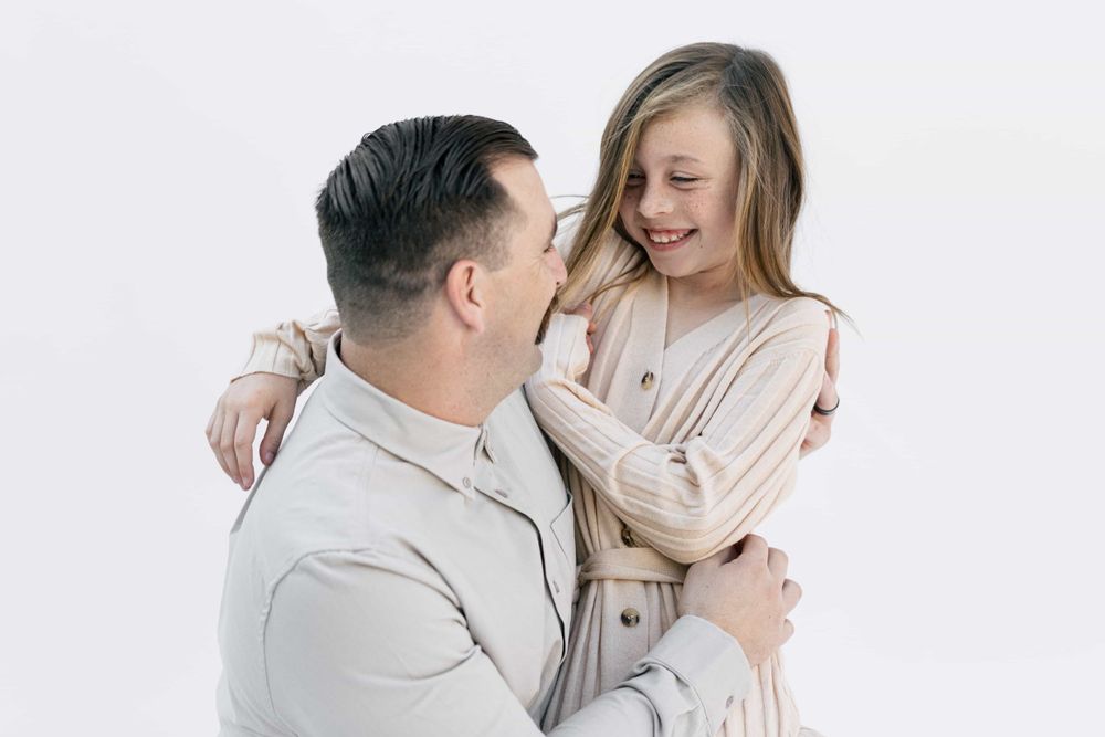 A father and daughter share a sweet embrace, smiling at each other against a white background.