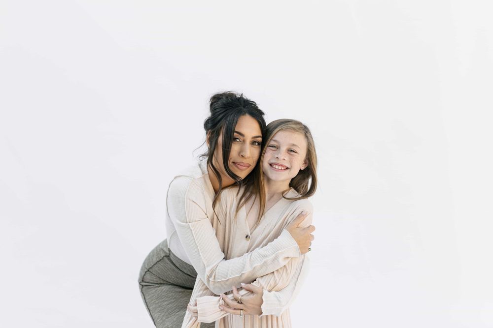 A mother and daughter share a warm hug, both smiling brightly against a white studio background.