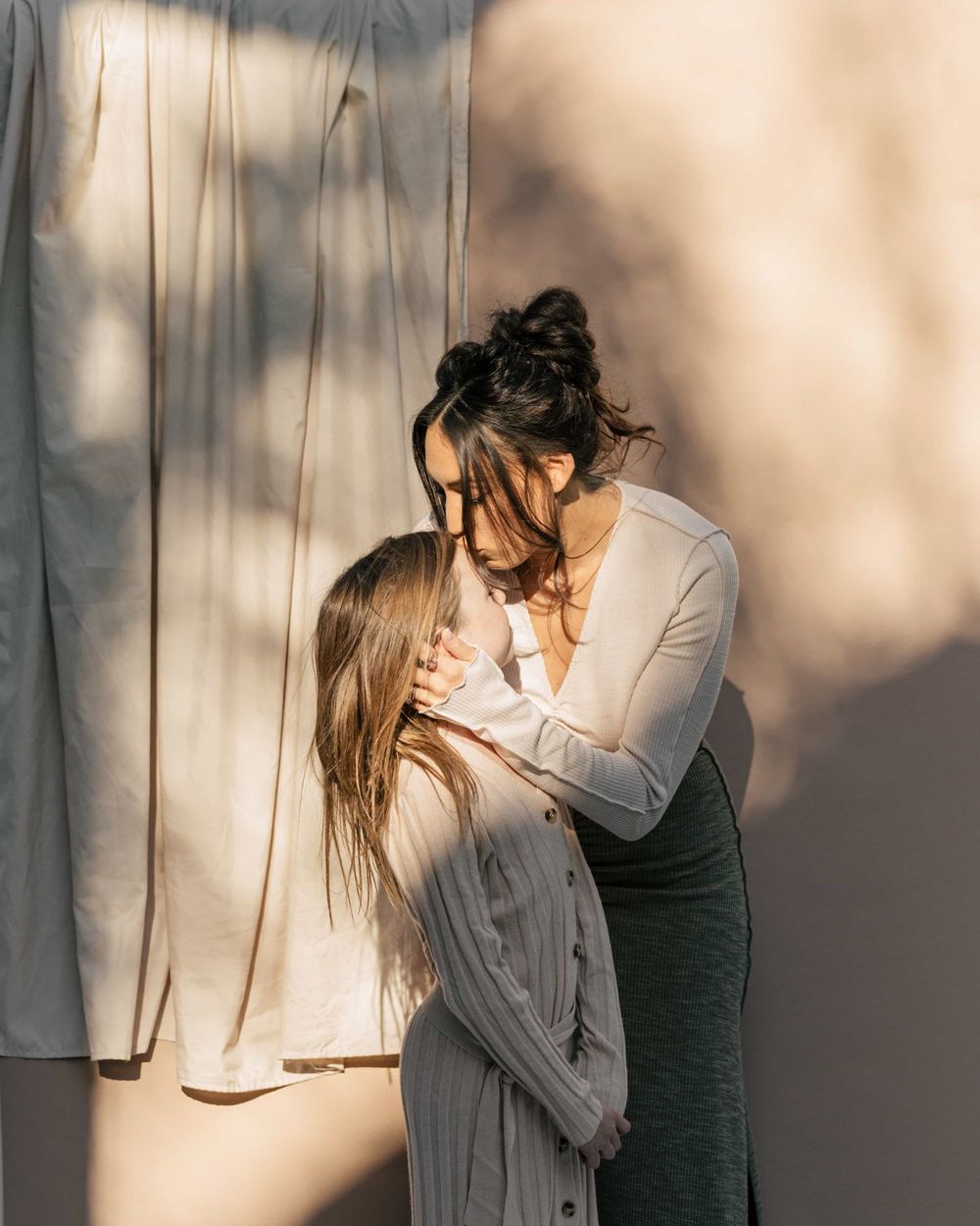 Mother tenderly embracing her young daughter against a sunlit neutral backdrop.