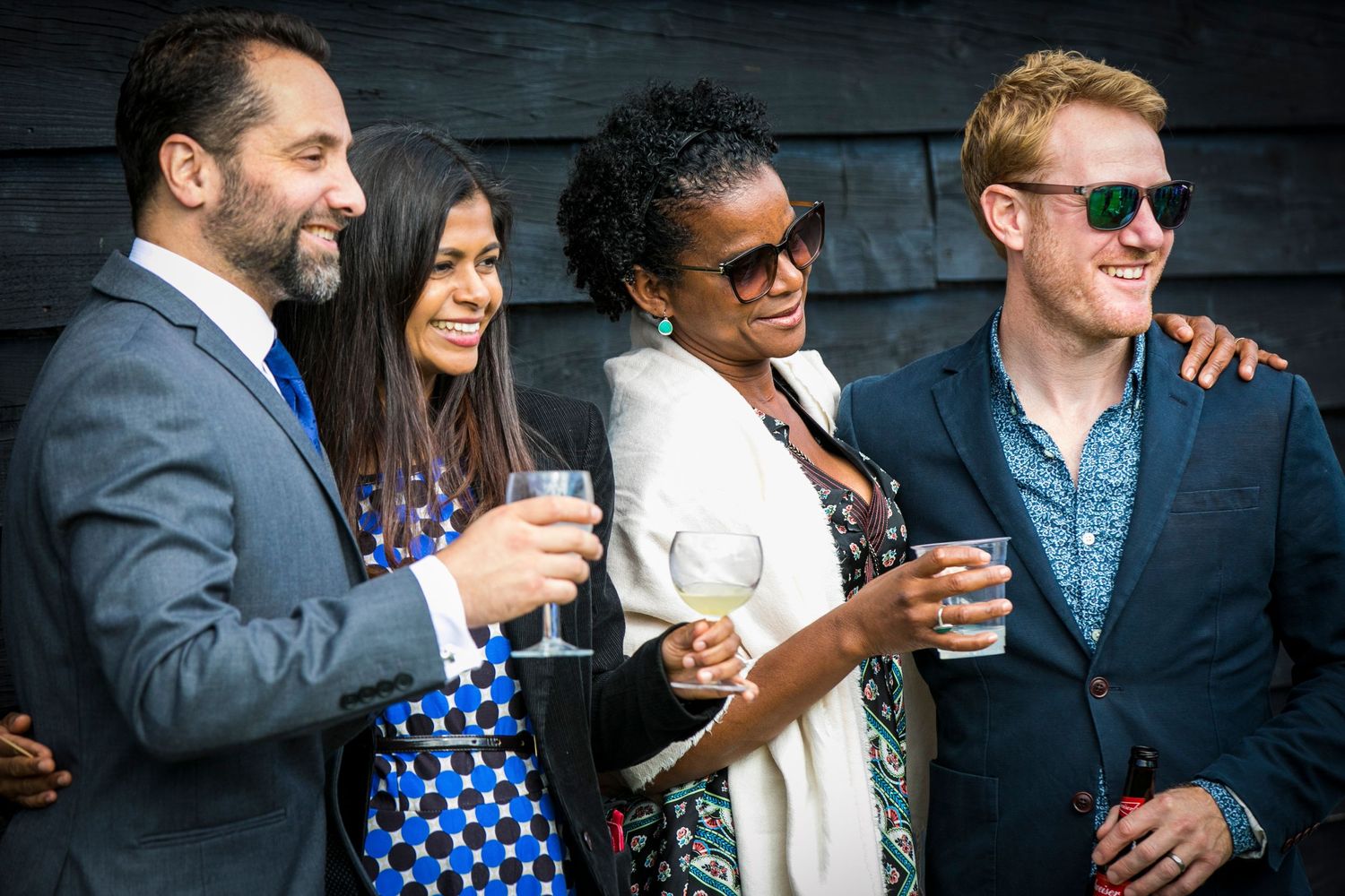 Wedding party socializing and enjoying beverages against dark wooden backdrop.