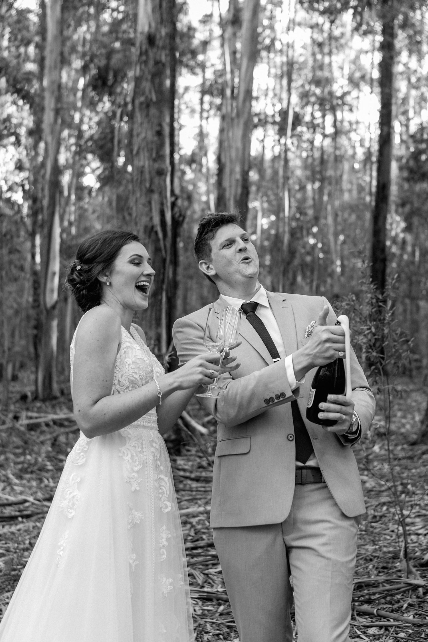A wedding couple laughs as the groom pops a bottle of champagne.