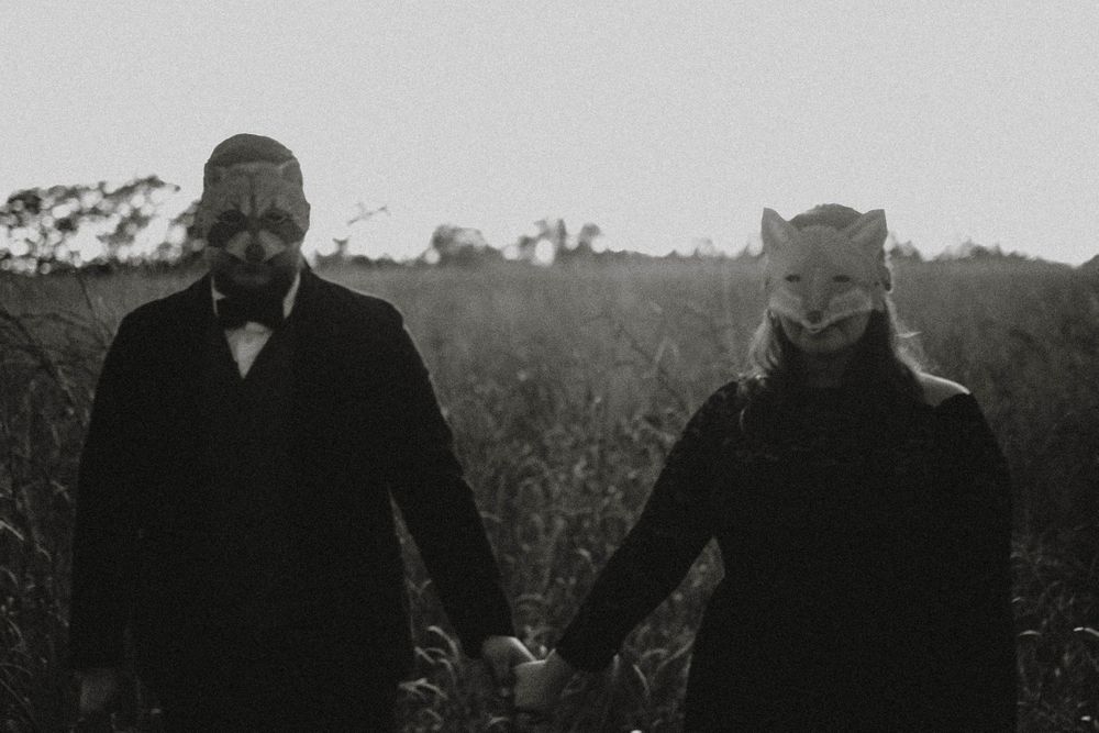 Two figures in dark clothing holding hands in a field, one wearing an eerie mask.