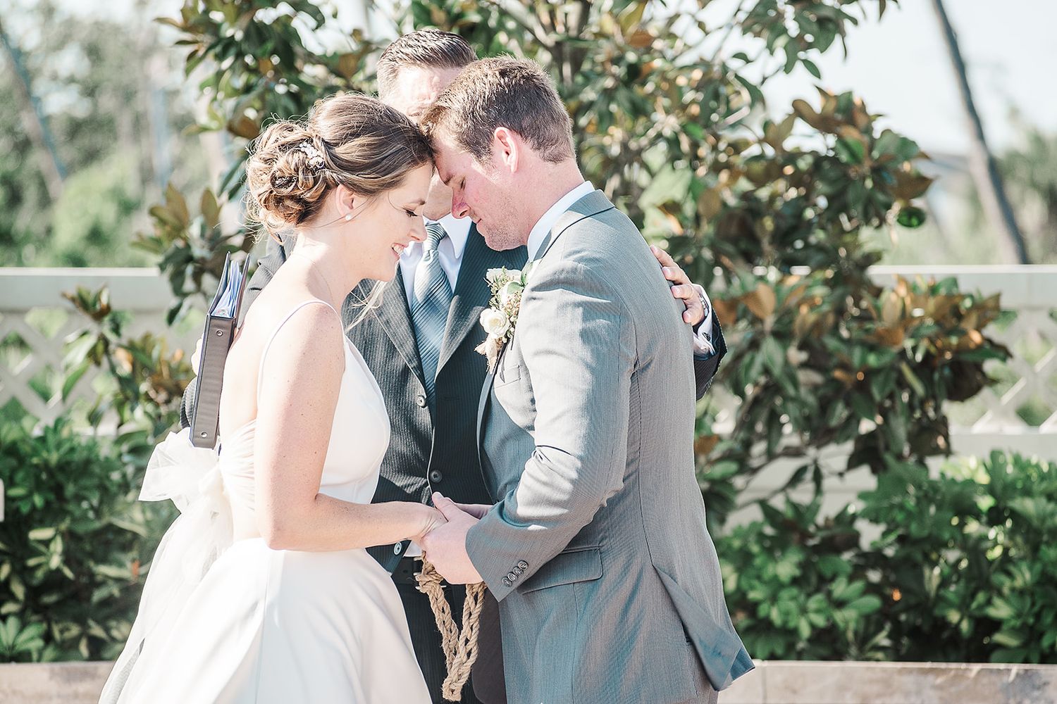 A bride and groom share an intimate moment during their sunny outdoor wedding ceremony surrounded by lush greenery.