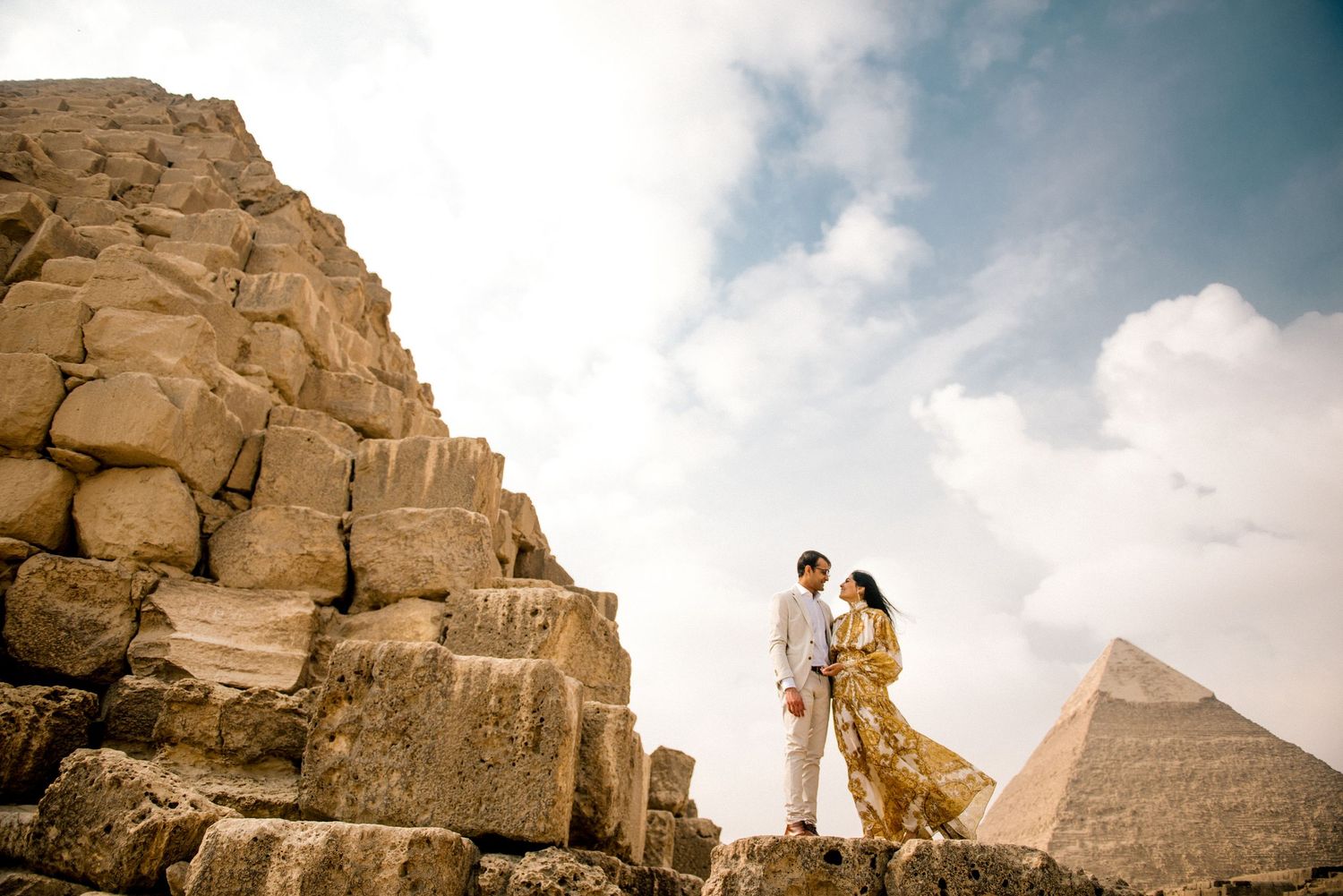 A couple stands together on ancient stone blocks with the iconic pyramids of Giza rising in the background.