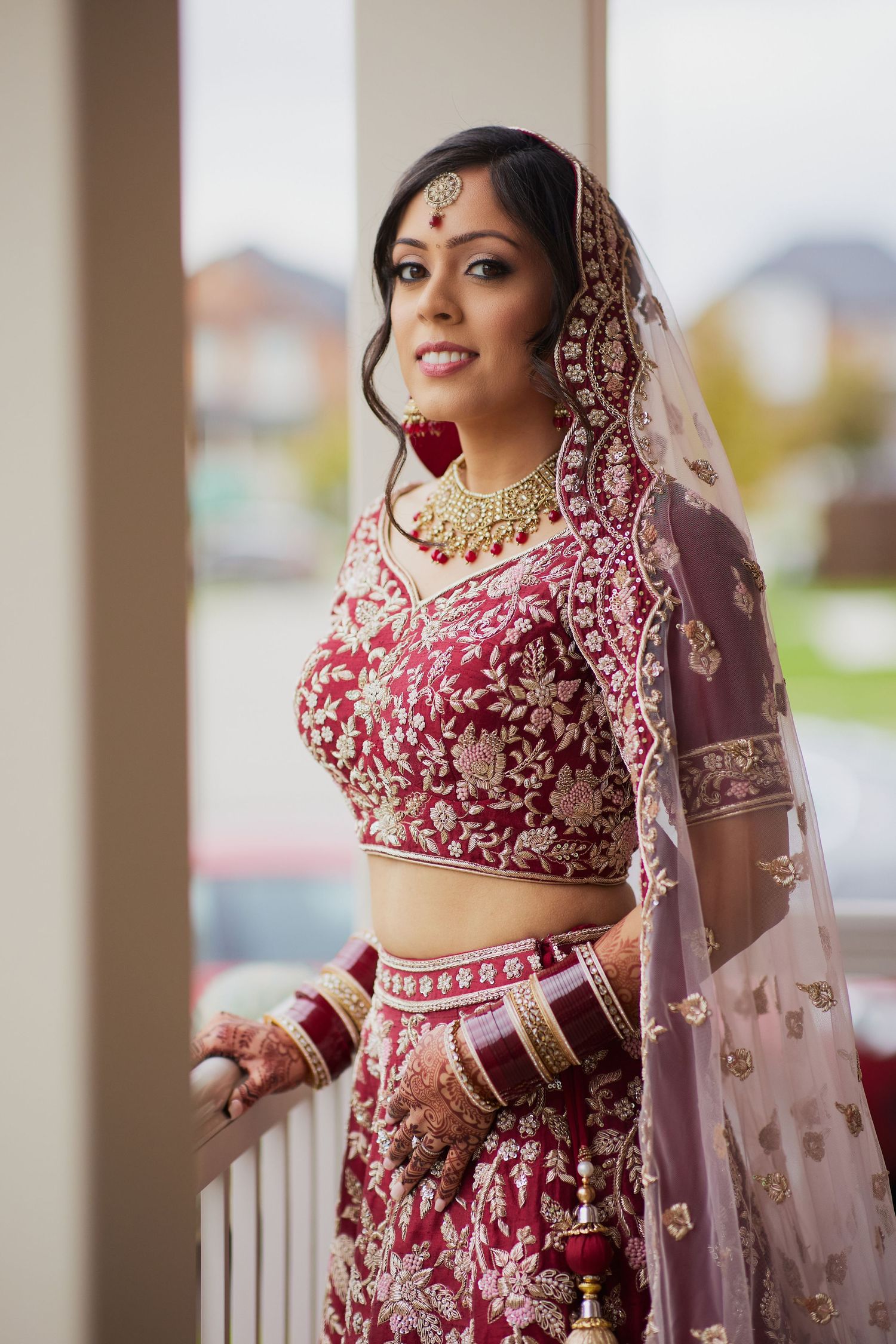 Traditional Indian bride in ornate red lehenga with embroidered details poses on porch.