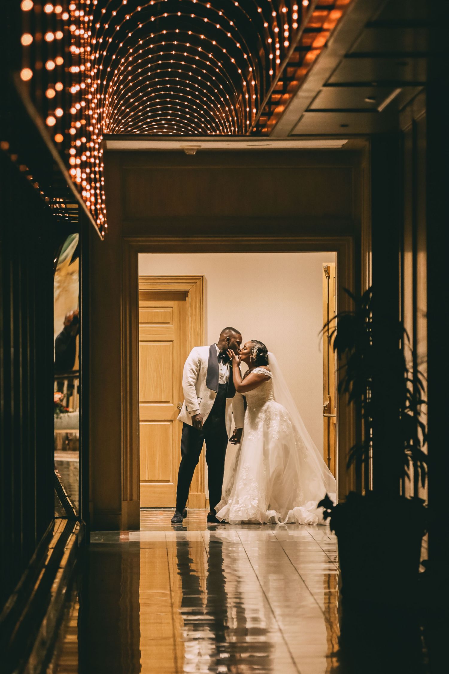 Newlyweds share a romantic kiss in a warmly lit hallway adorned with a stunning circular ceiling light display.