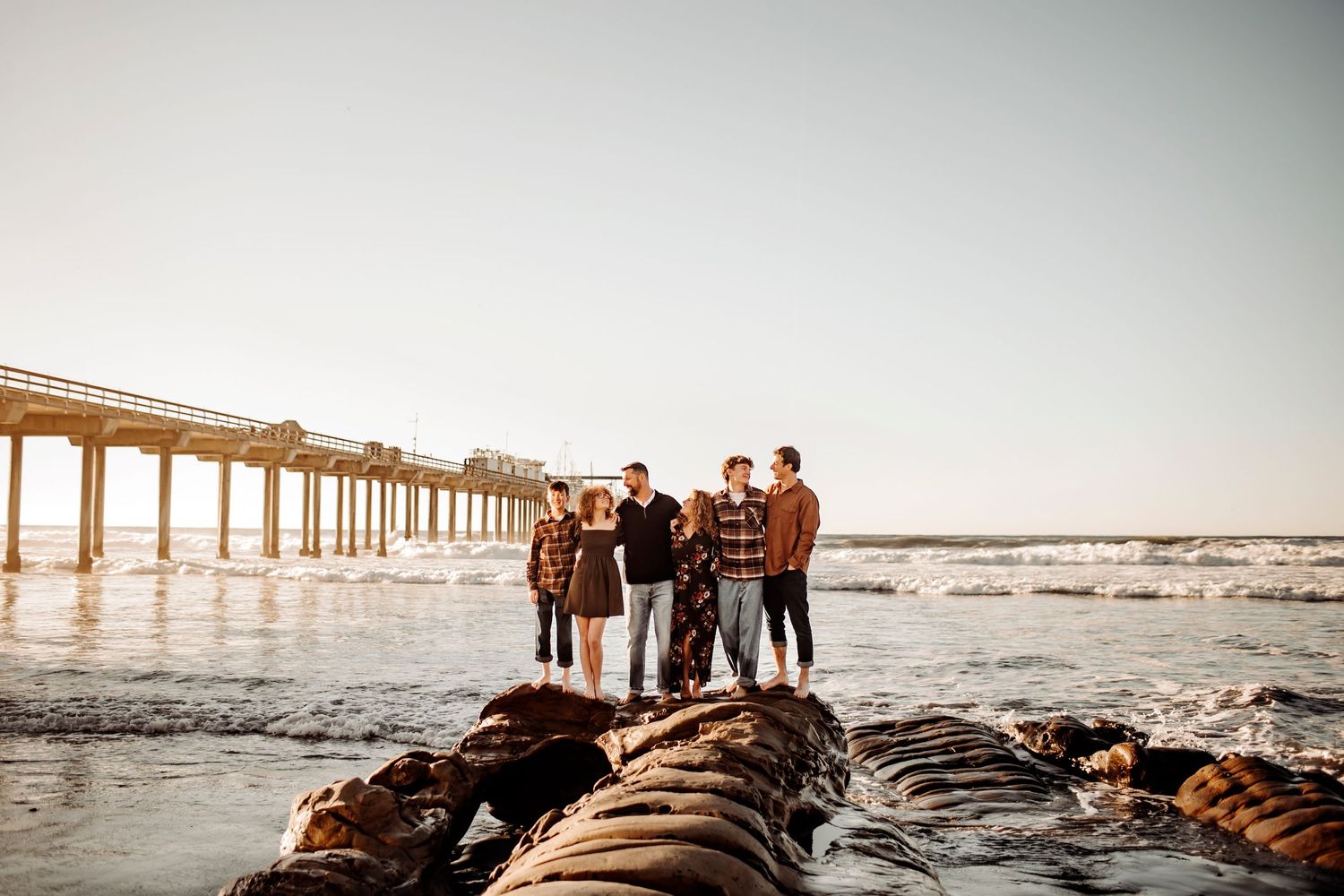 A family of six posing together on rocks near a pier at the beach, smiling warmly with waves and a serene ocean backdrop.