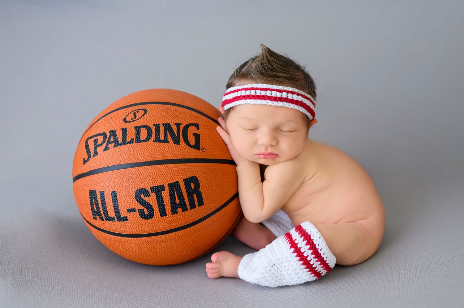 Baby in athletic sweatband and socks poses with orange Spalding All-Star basketball on gray background.