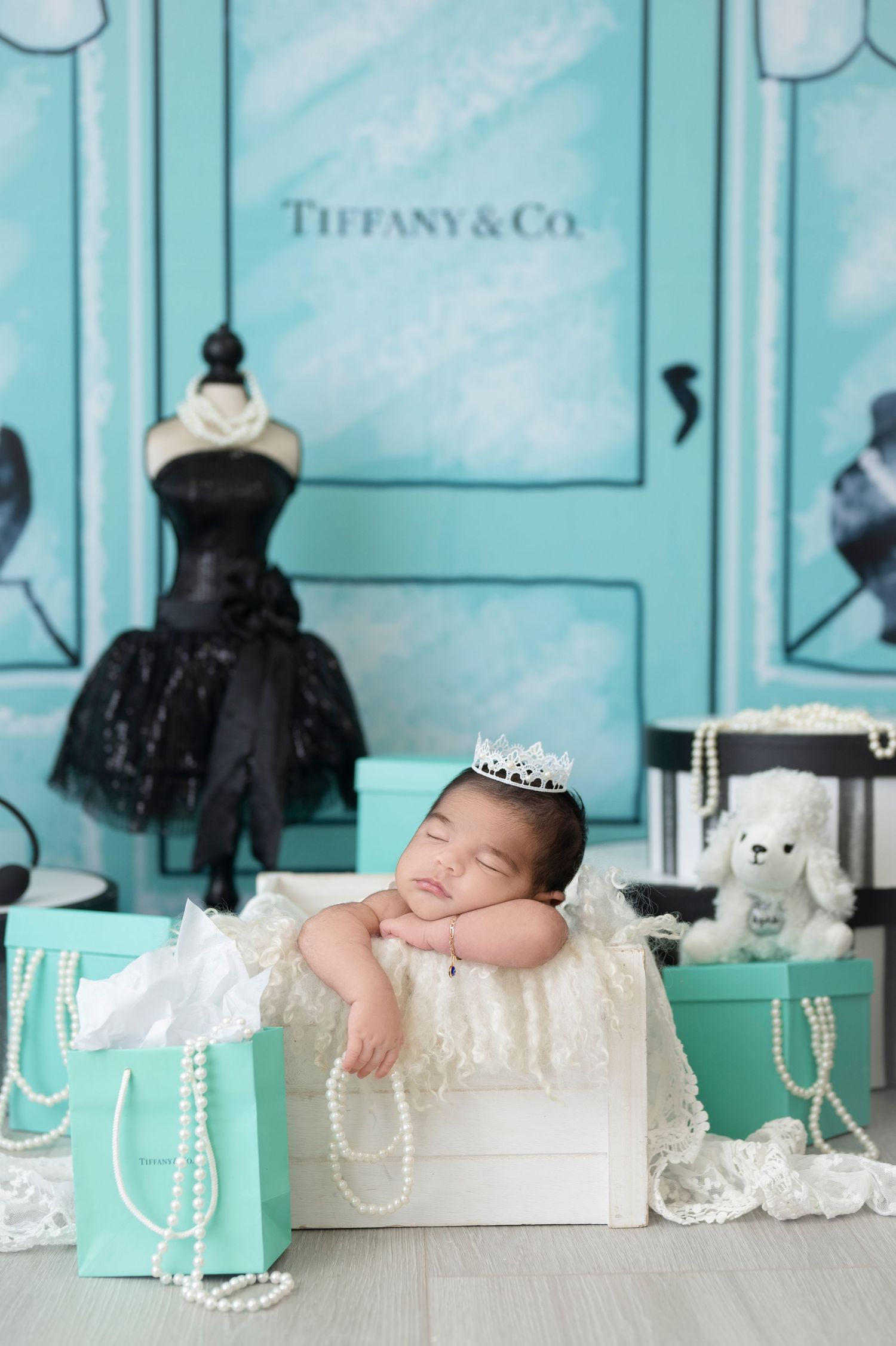 A newborn sleeps on a Tiffany blue gift box surrounded by pearls and shopping bags in an upscale boutique setting.