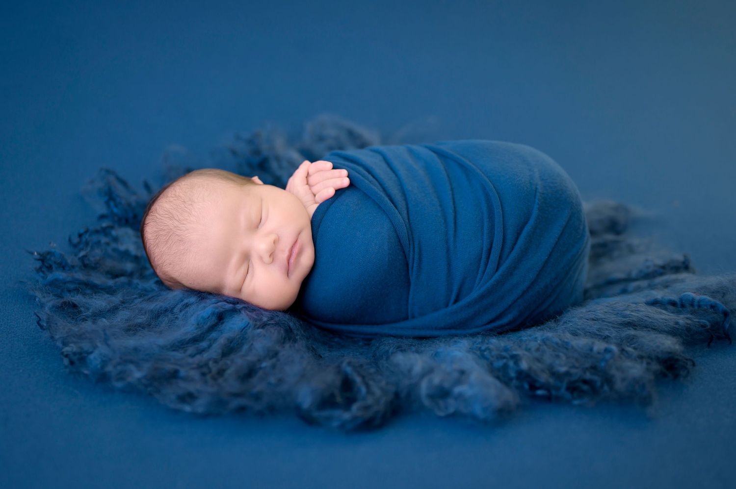 A sleeping newborn baby wrapped in blue fabric lies on a fluffy navy blue blanket with blue background blanket.
