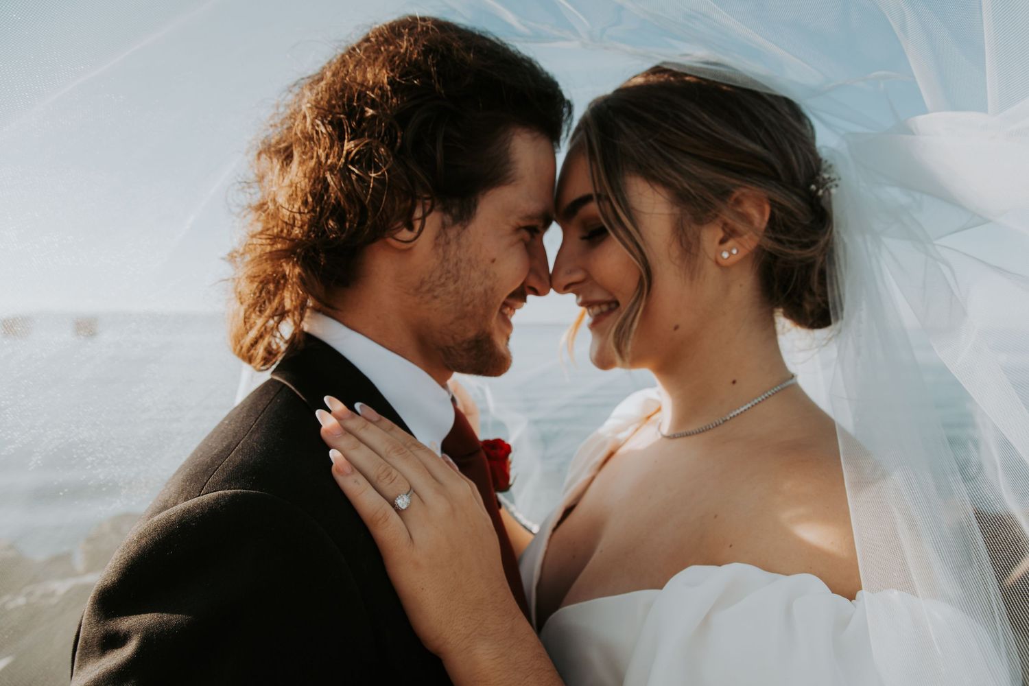Bride and groom touch foreheads and smile as a veil blows around them during a sunny seaside wedding portrait.