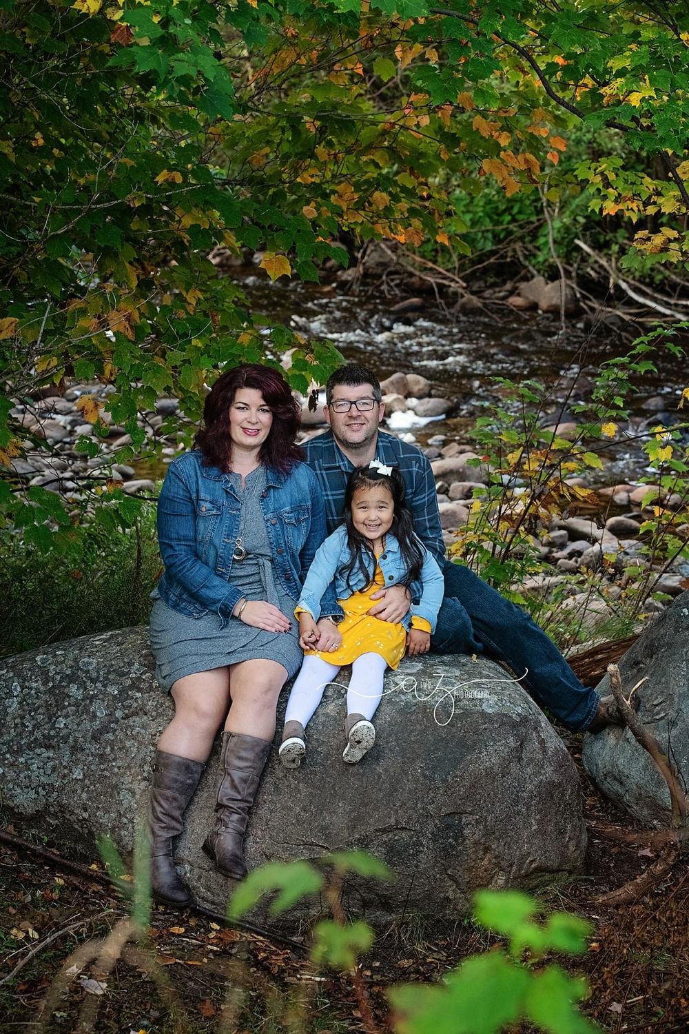 Family sits together on large rock near flowing stream with autumn foliage overhead.