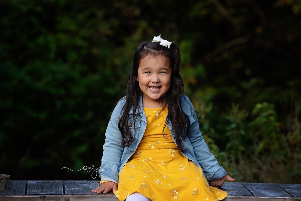 Child wearing yellow dress and denim jacket sits joyfully on wooden surface against blurred forest background.
