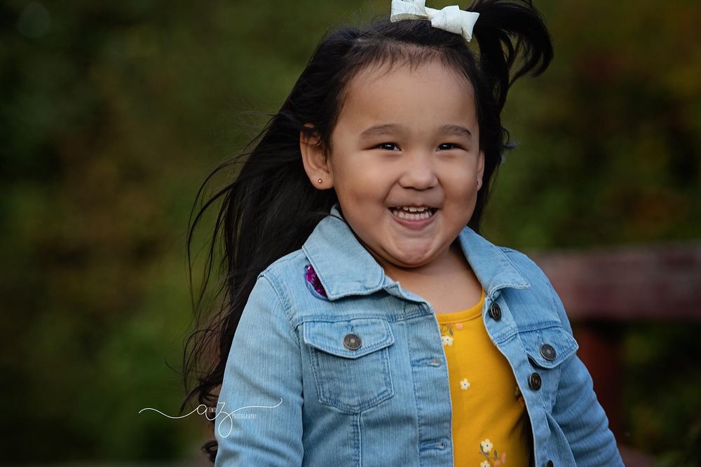 Child in denim jacket and yellow dress smiles brightly with windblown hair outdoors.