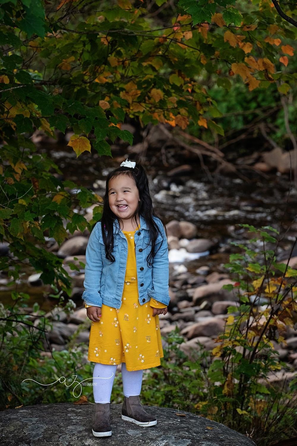 Child in bright yellow dress poses near rocky stream with autumn leaves overhead.