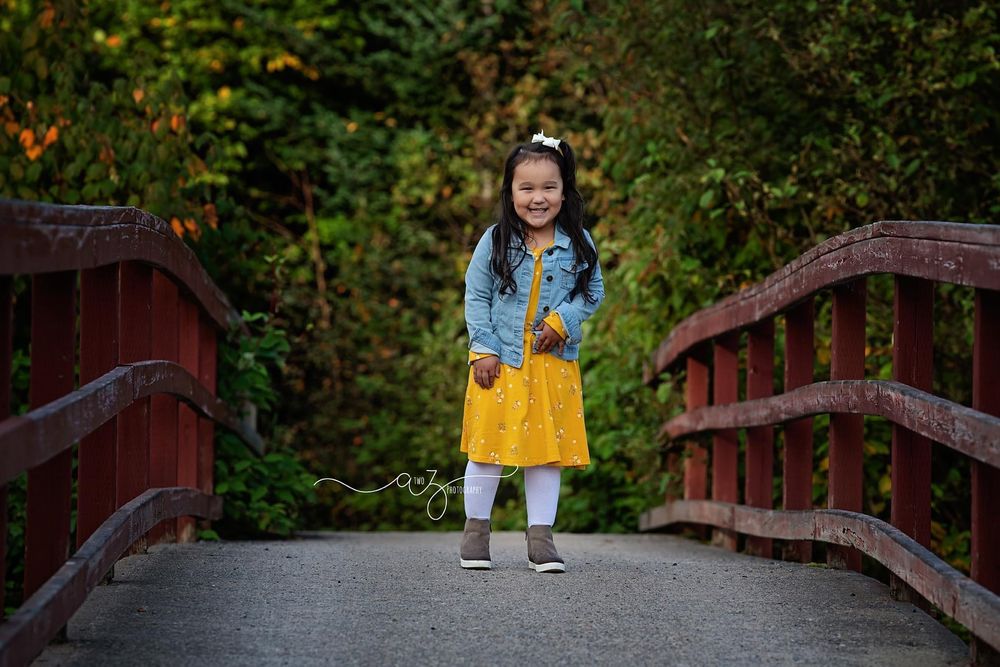 Child in yellow dress stands on red wooden bridge surrounded by autumn foliage.