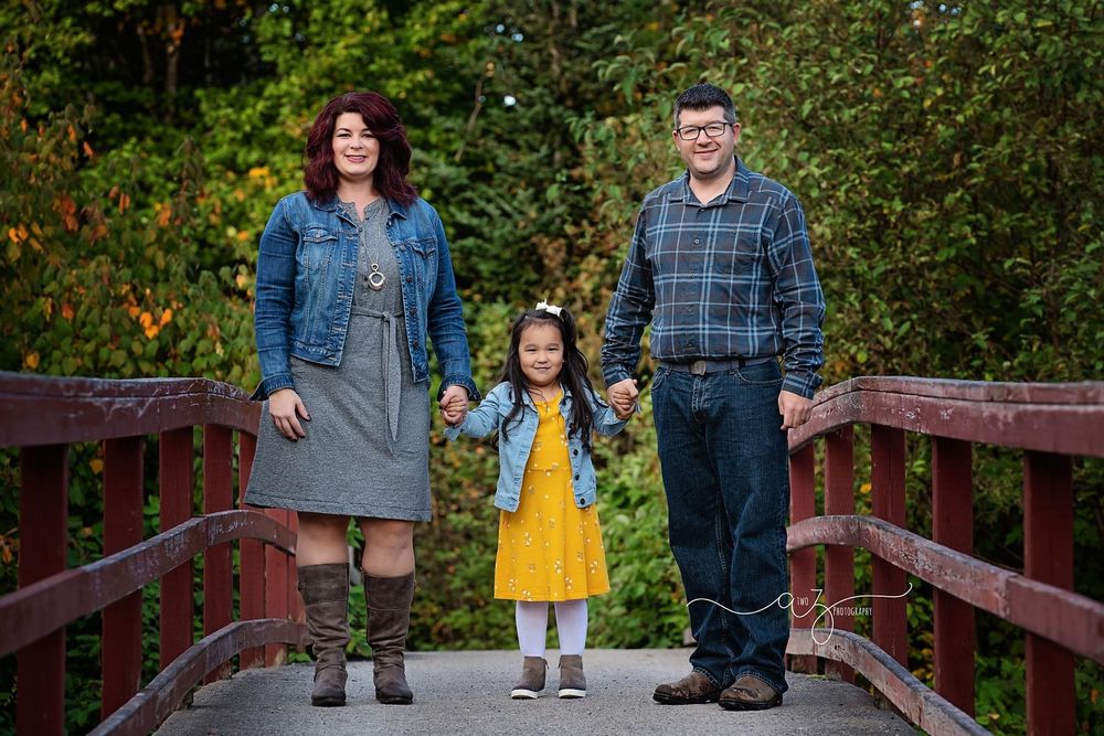 Family stands together on wooden bridge with natural fall foliage in background.