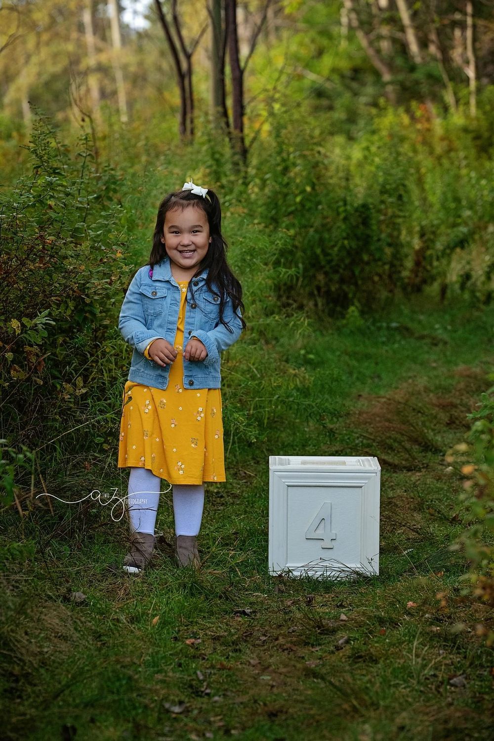 Child in yellow dress and denim jacket stands by white milestone marker on forest trail.