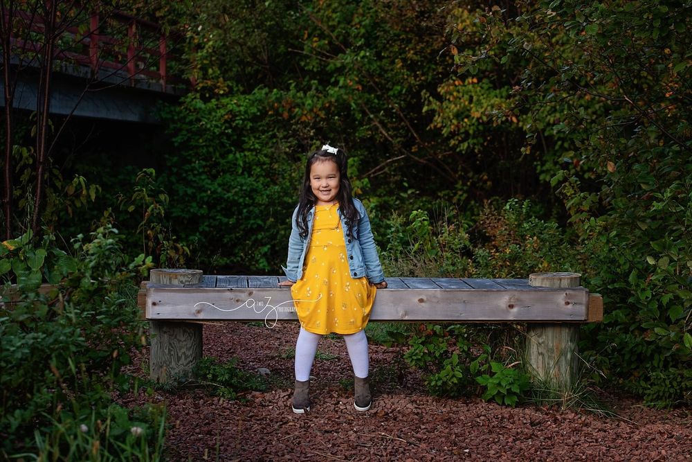 Child in yellow dress and denim jacket sits on wooden bench surrounded by greenery.