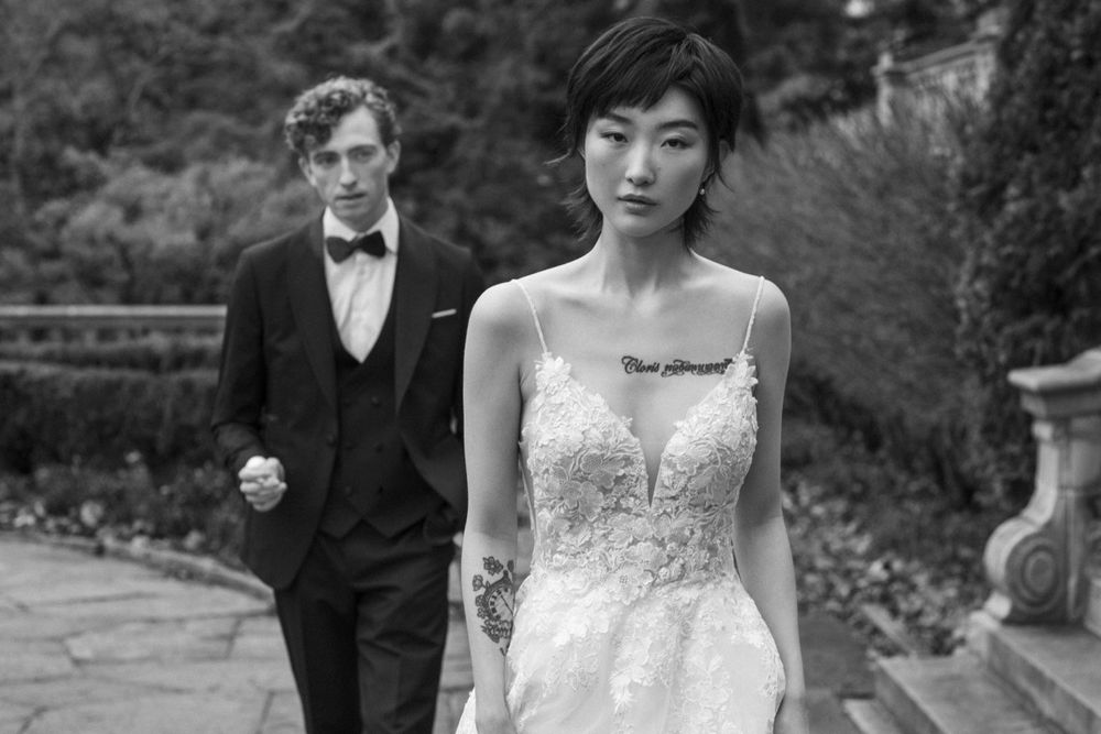 A wedding couple poses in formal attire on a stone path surrounded by greenery.