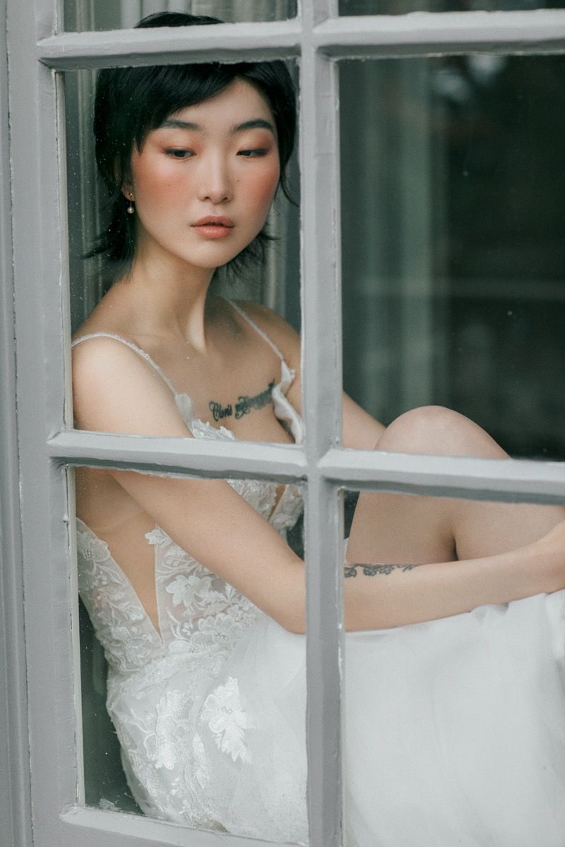 A bride in a delicate lace wedding dress poses behind a window frame with natural lighting.