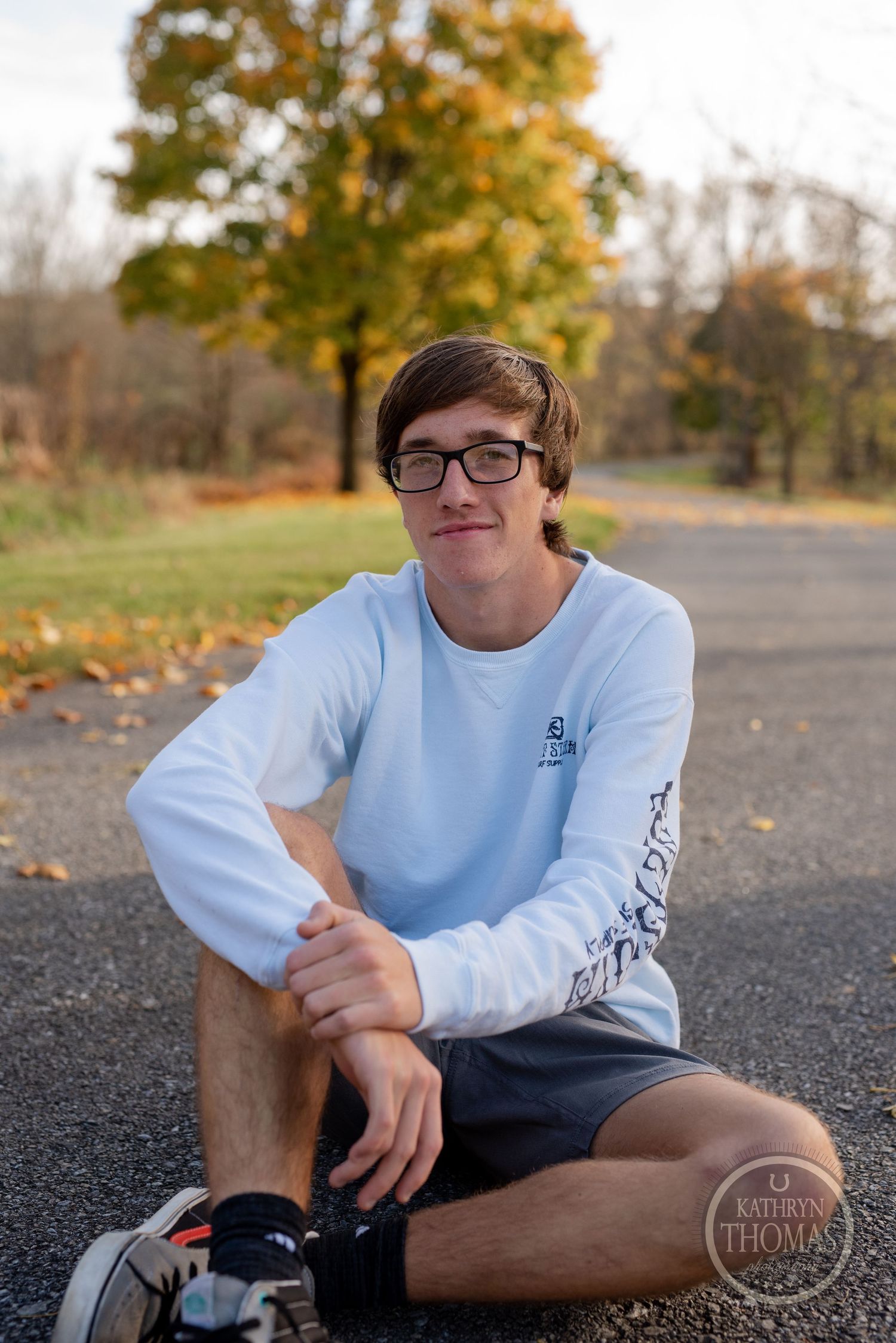Person in white long-sleeve shirt sits casually on ground during autumn photoshoot.