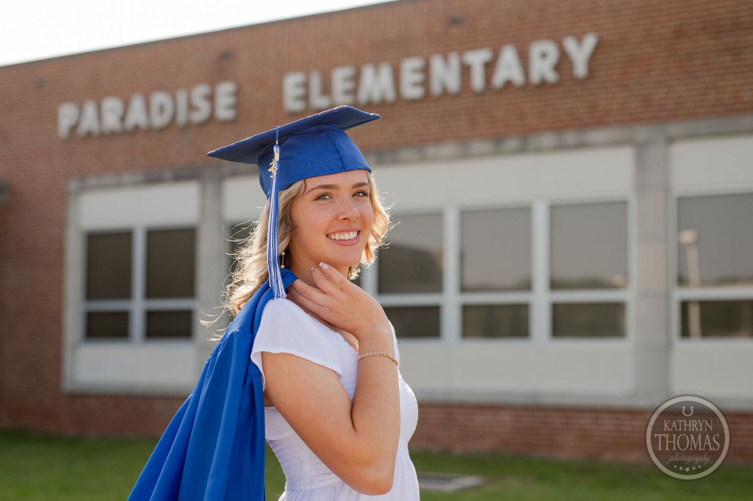 Graduate in blue cap and gown smiles joyfully outside Paradise Elementary.