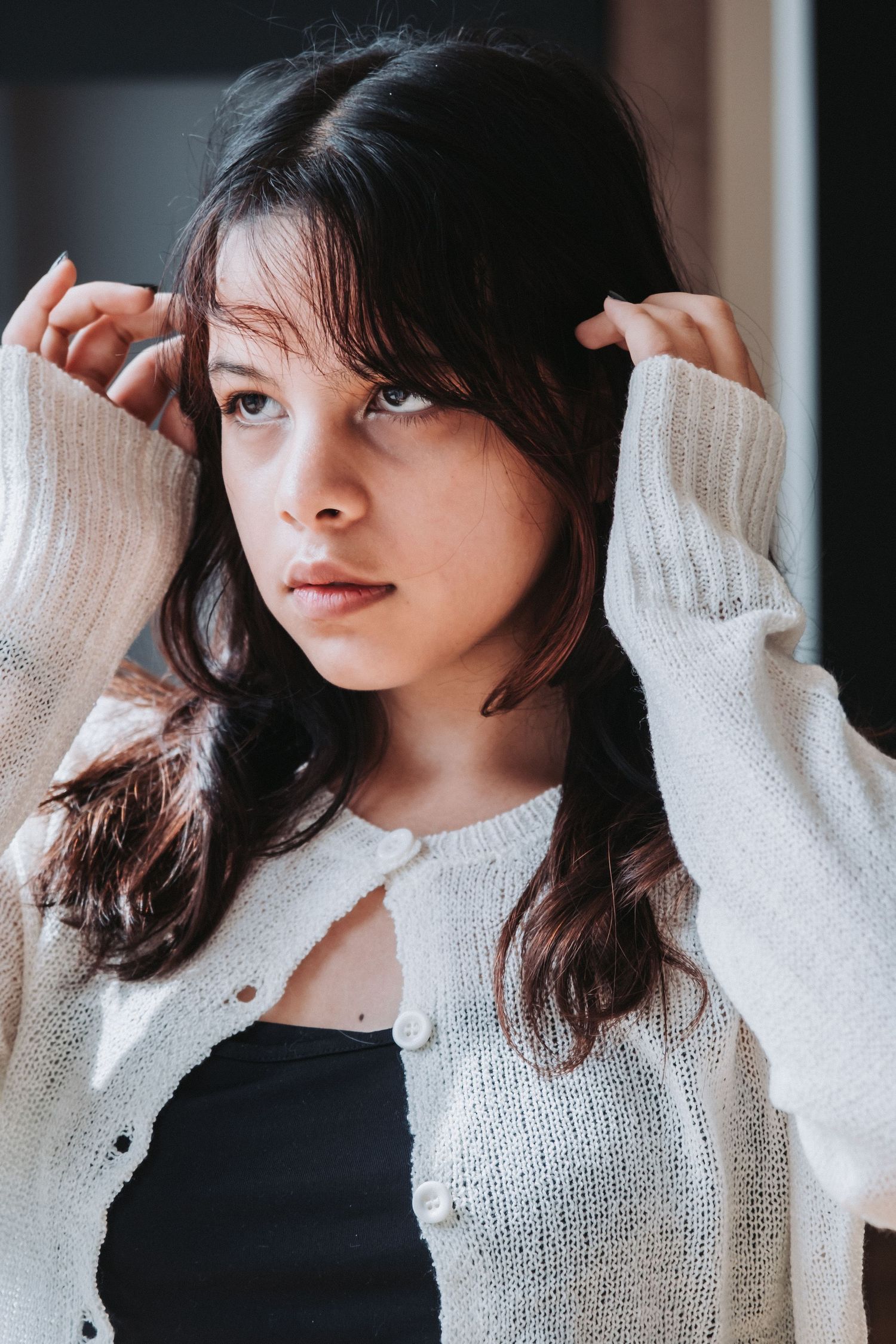 Woman in white sweater adjusting her hair in moody indoor portrait.