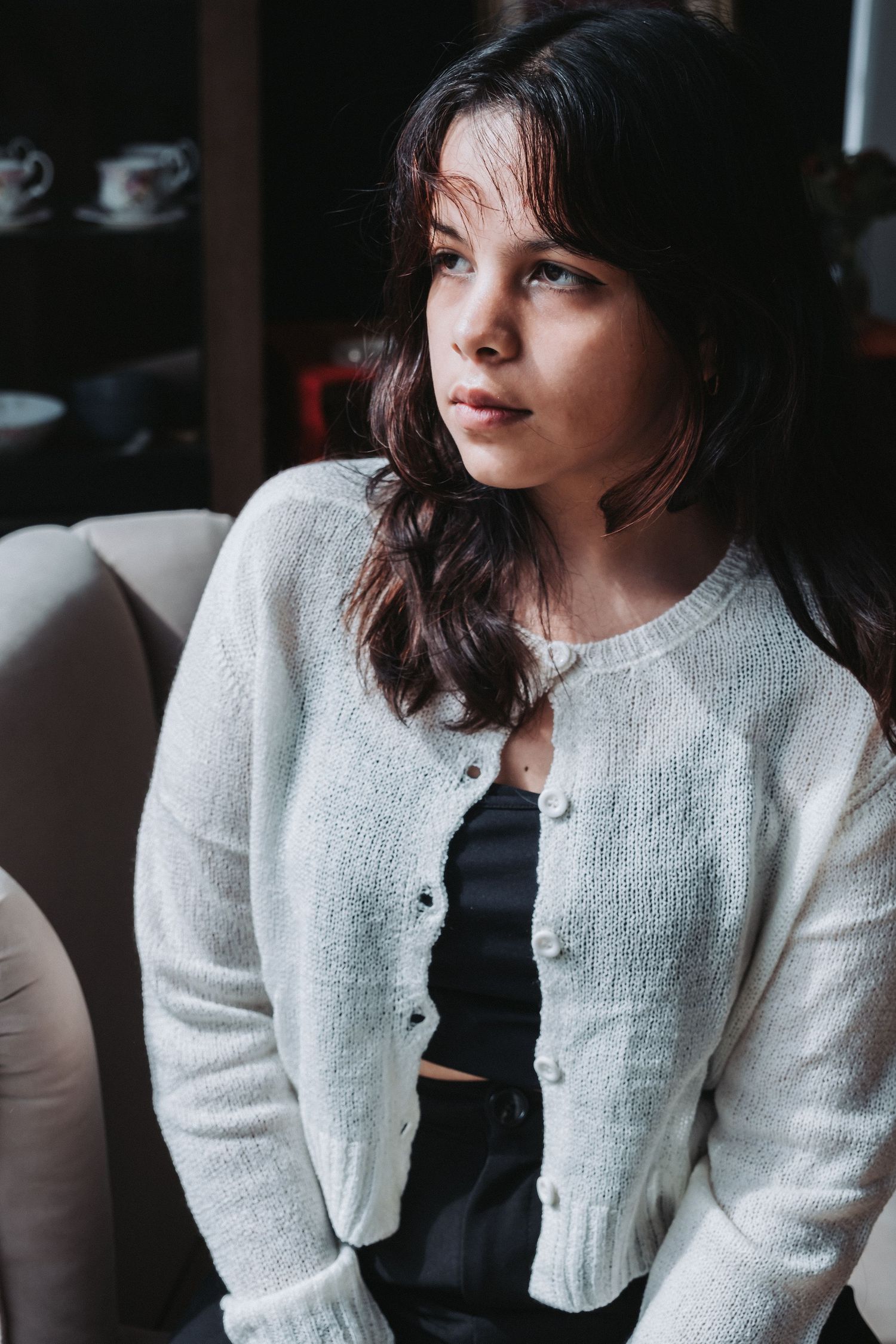 Woman in white cardigan and dark top poses by window in casual indoor portrait.
