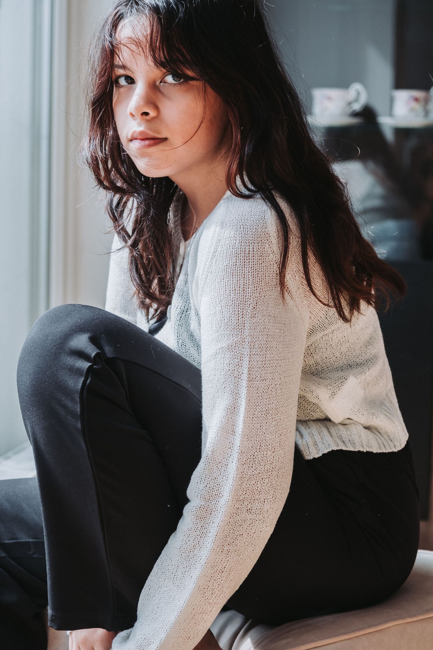Woman in white sweater and dark pants sitting casually by window in moody indoor portrait.